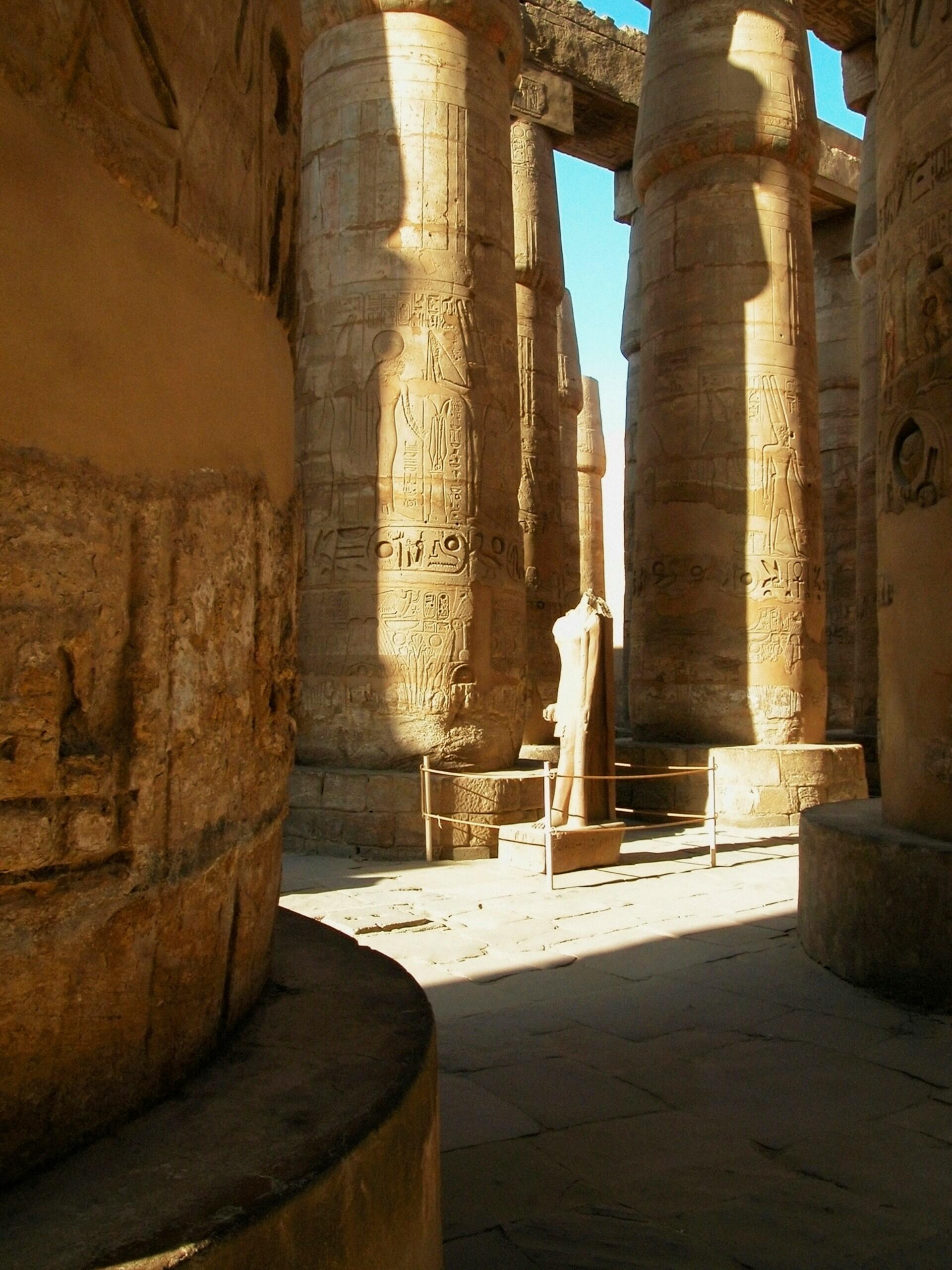 Ancient Egyptian temple with columns covered in hieroglyphs, sunlit pathway leading to a statue.