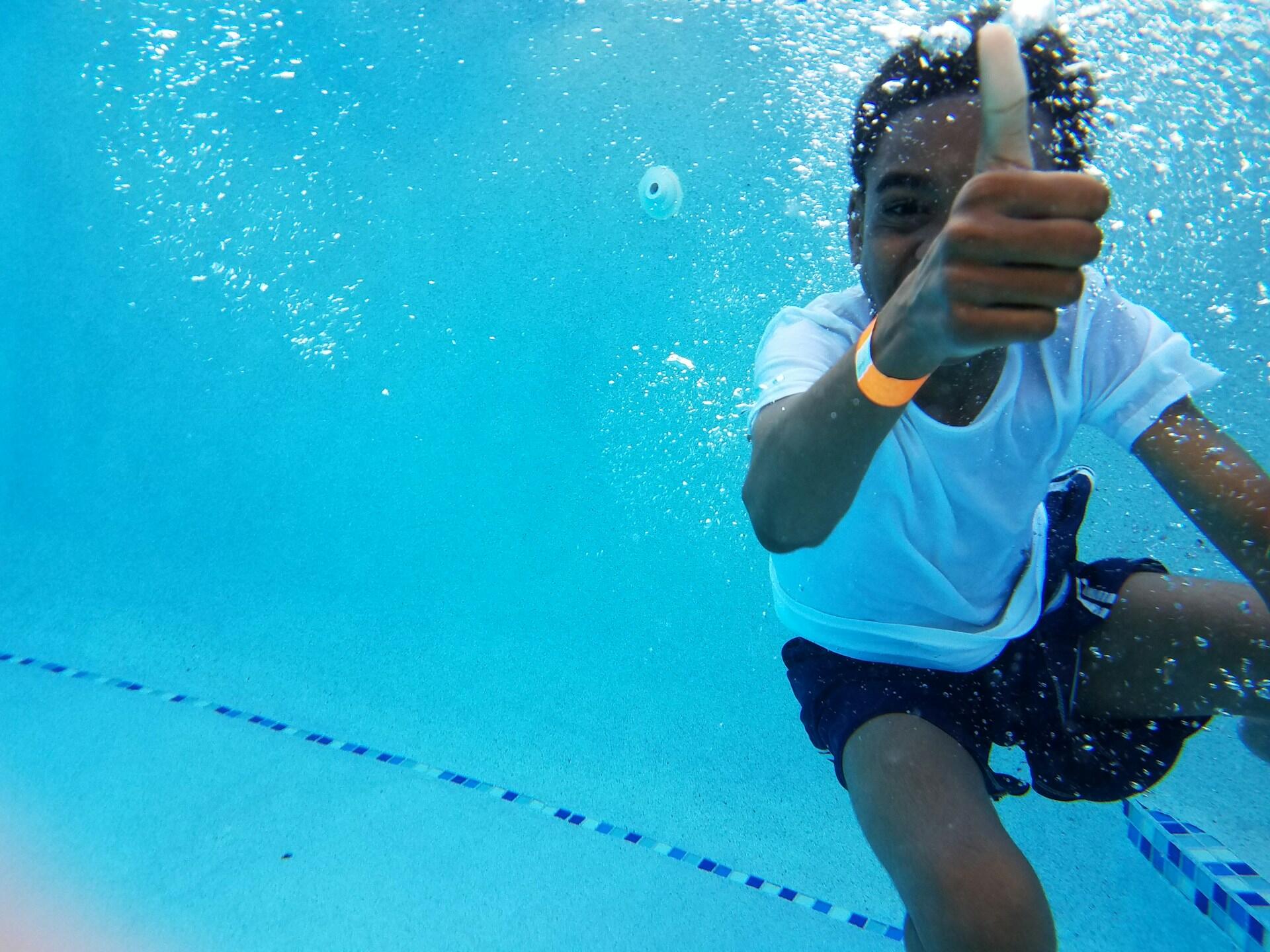 A child wearing a white tee shirt underwater, showing thumbs up.