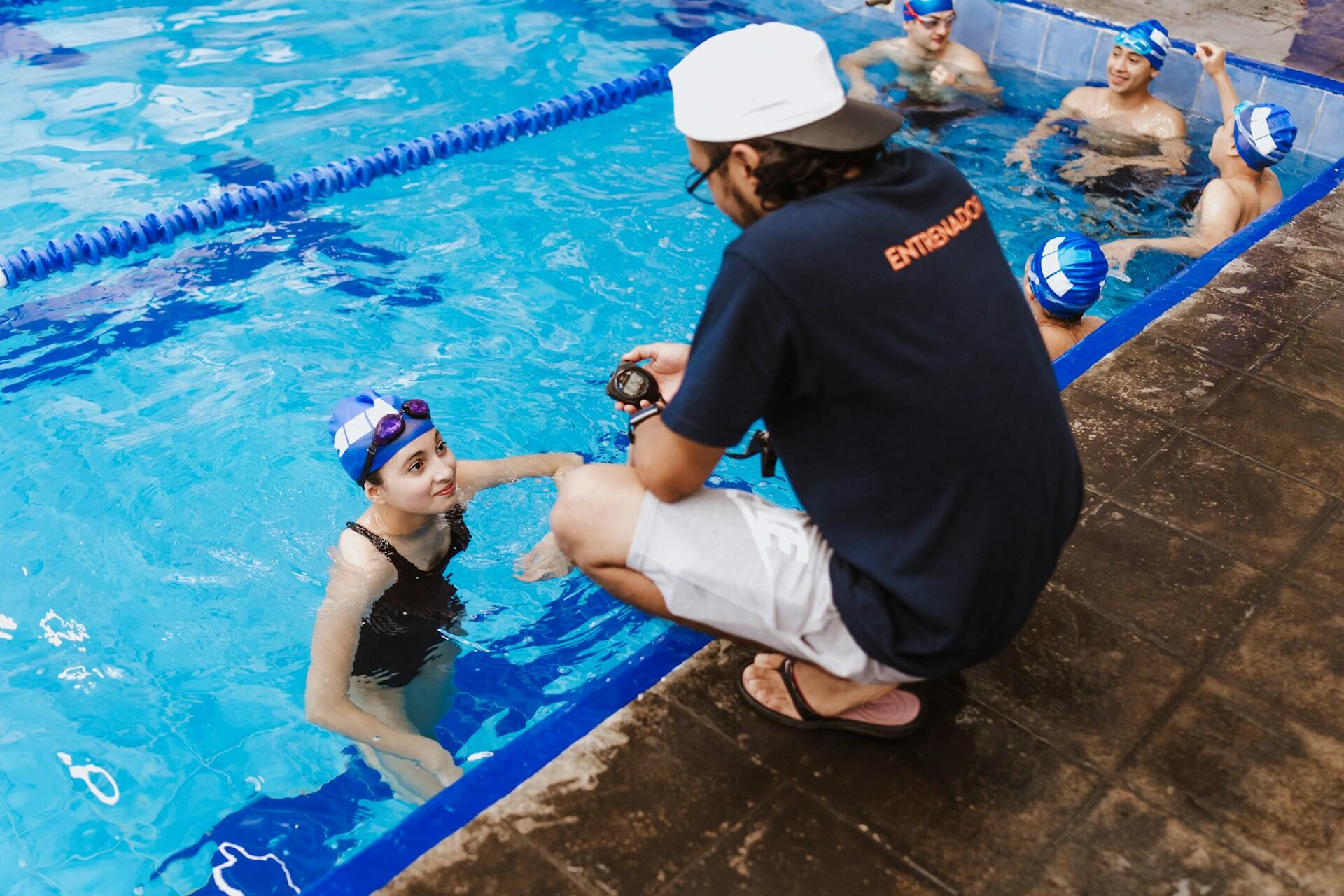 A man in a dark tee shirt squats poolside to instruct a swimmer.