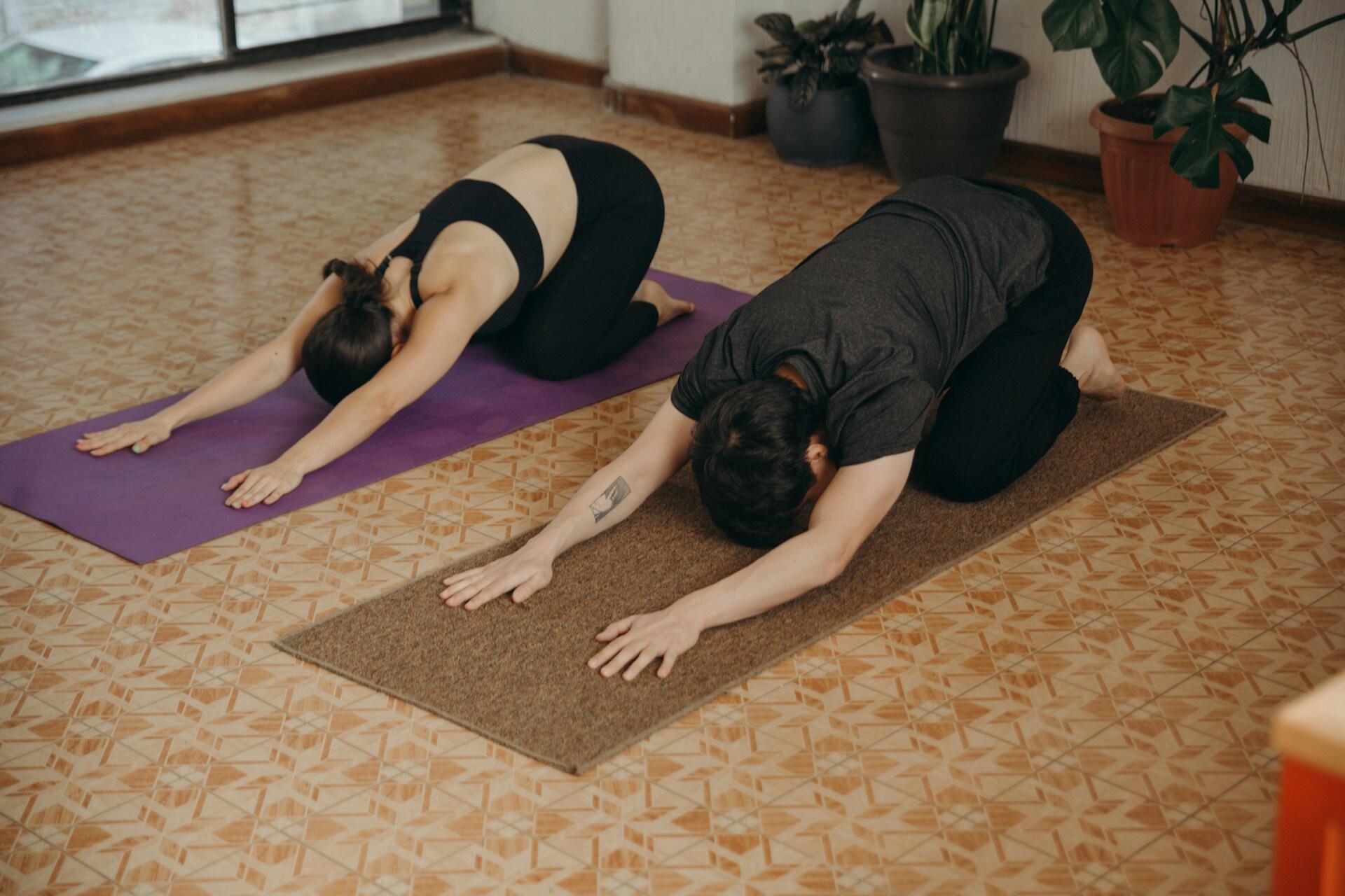 Two people practicing yoga child's pose on mats indoors.