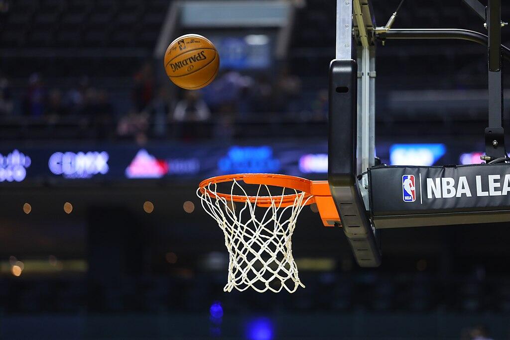 A basketball is seen mid-air above an NBA hoop, with the net and backboard visible in a stadium setting.