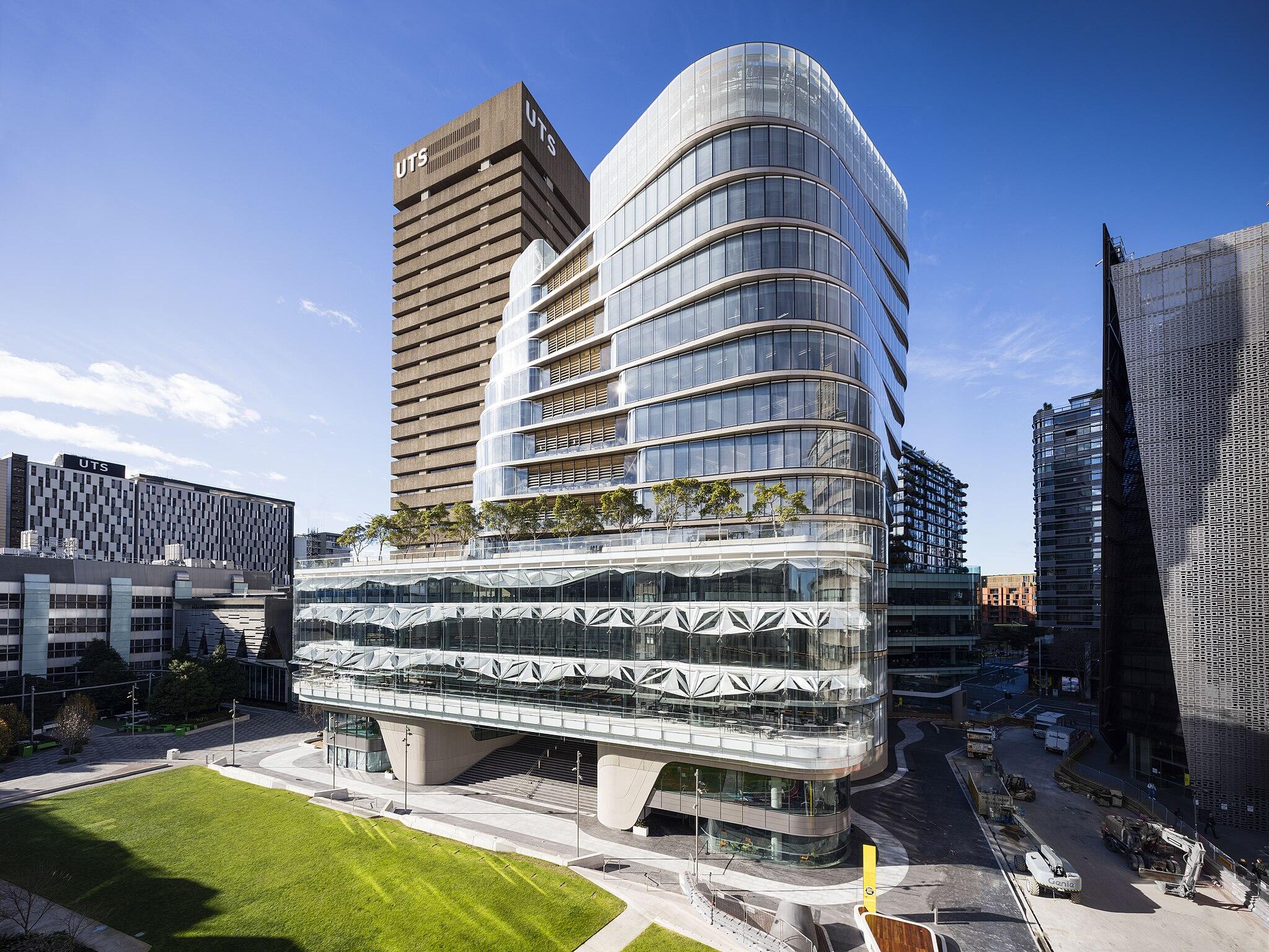 Modern UTS campus buildings, featuring a sleek glass structure with greenery, alongside a taller brick building against a clear blue sky.