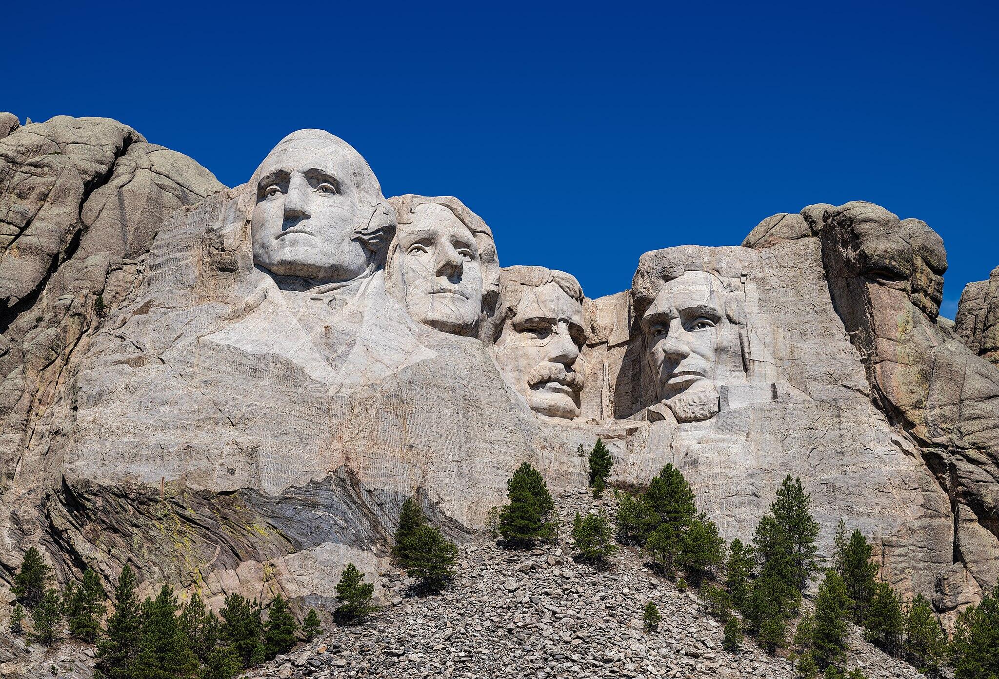 Majestic rock formations under a clear blue sky, with greenery at the base and hints of carved features on the cliffs above.