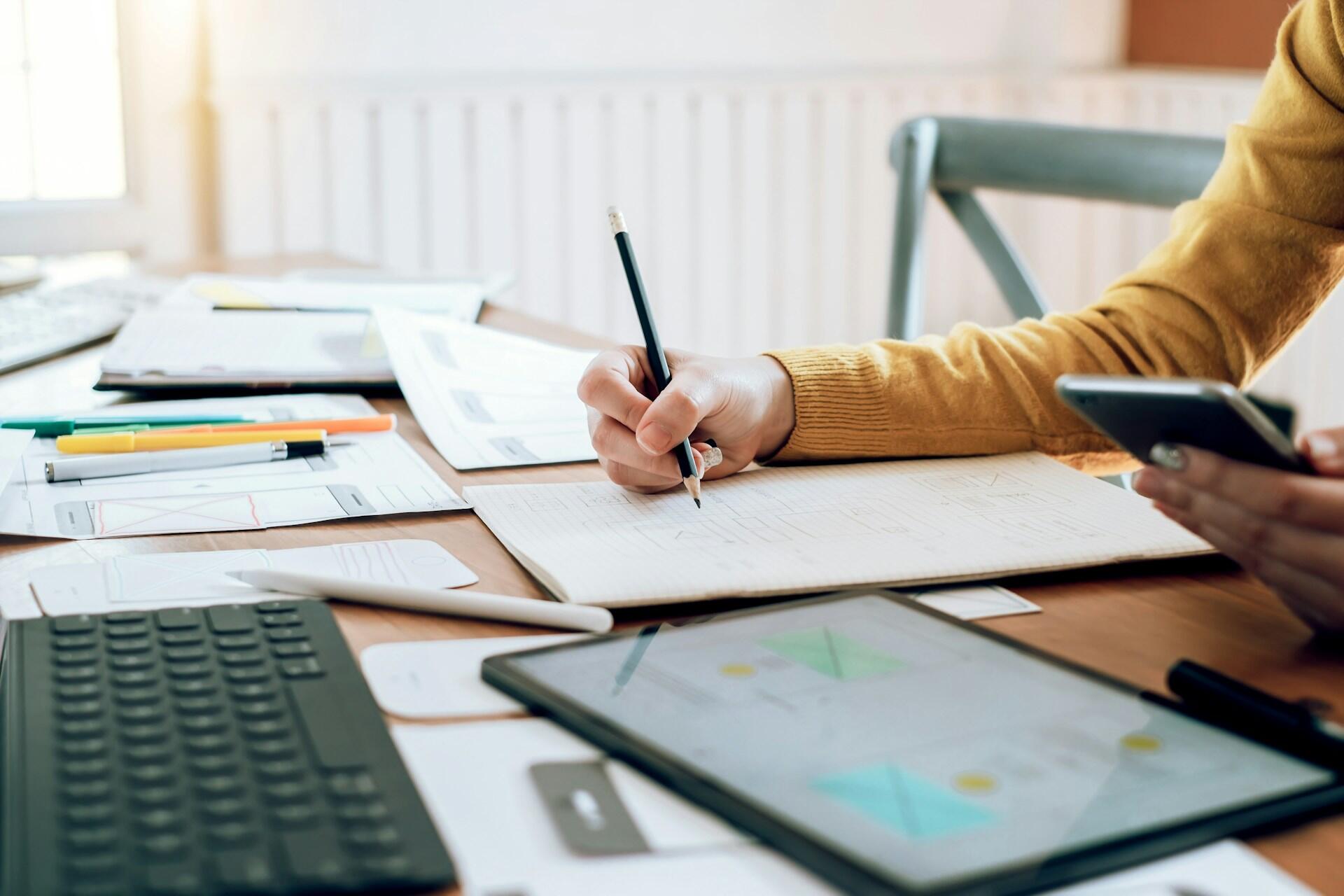 A person wearing a yellow jumper writes n a notepad, surrounded by electronic equipment.