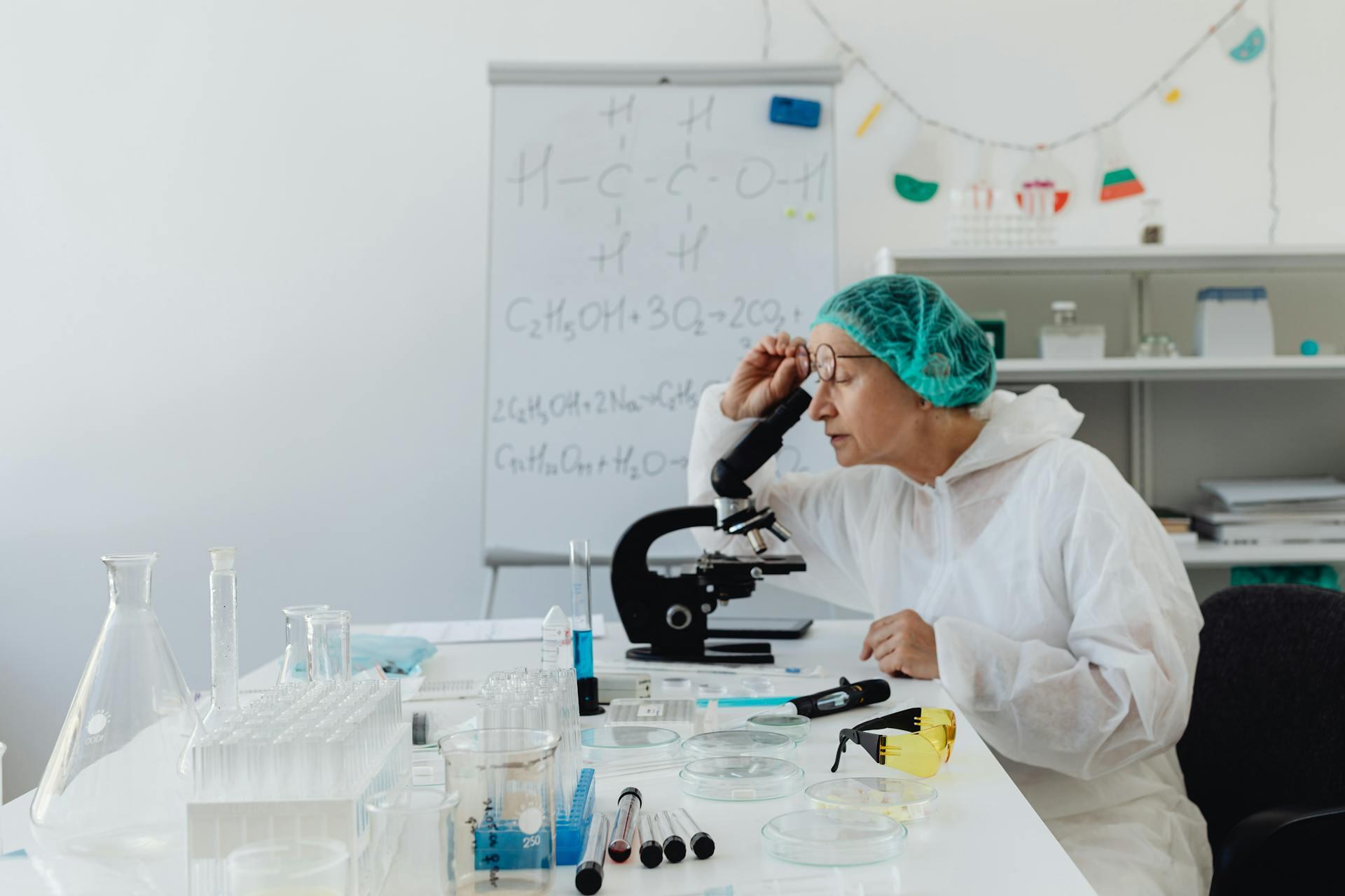 a chemist in a lab looking through a microscope, chemistry equations are written on the board in the back of the room