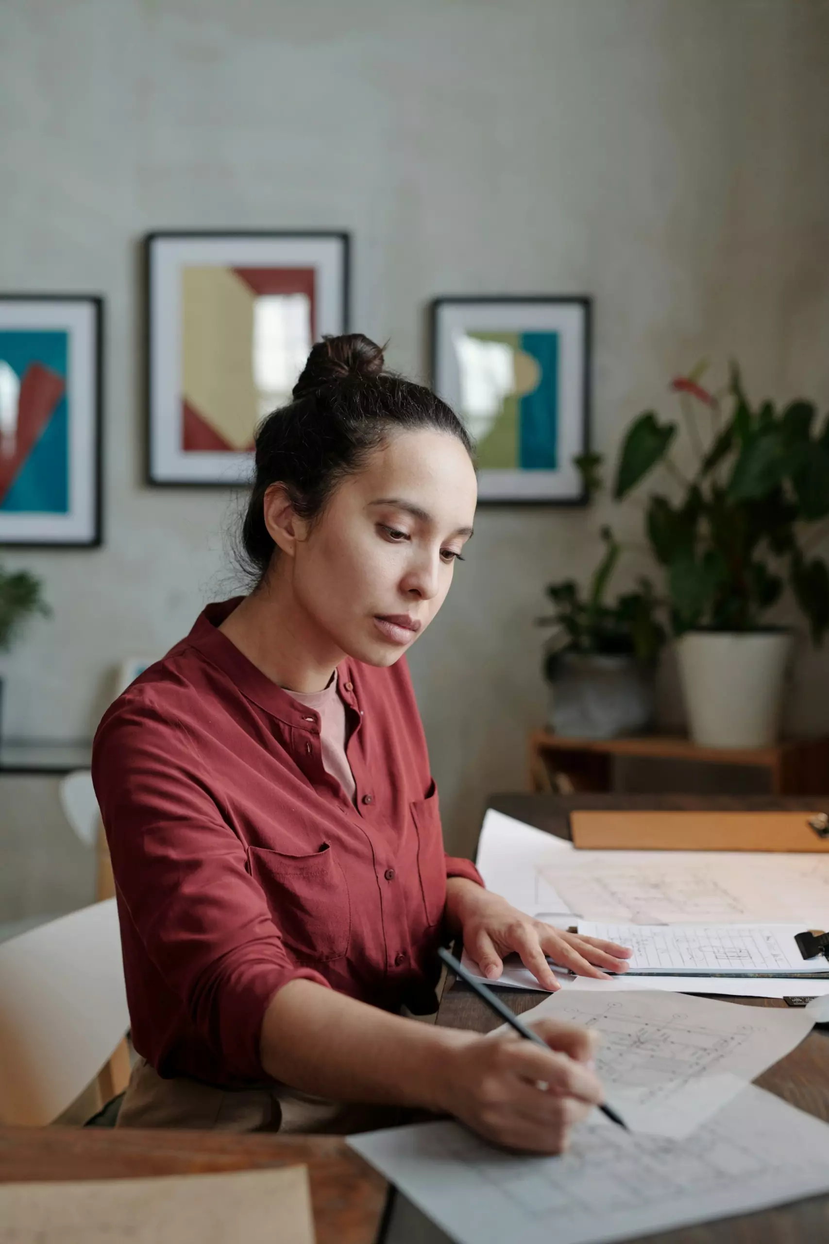 A person in a red shirt sketches plans at a wooden table, surrounded by greenery and framed artwork on the walls.
