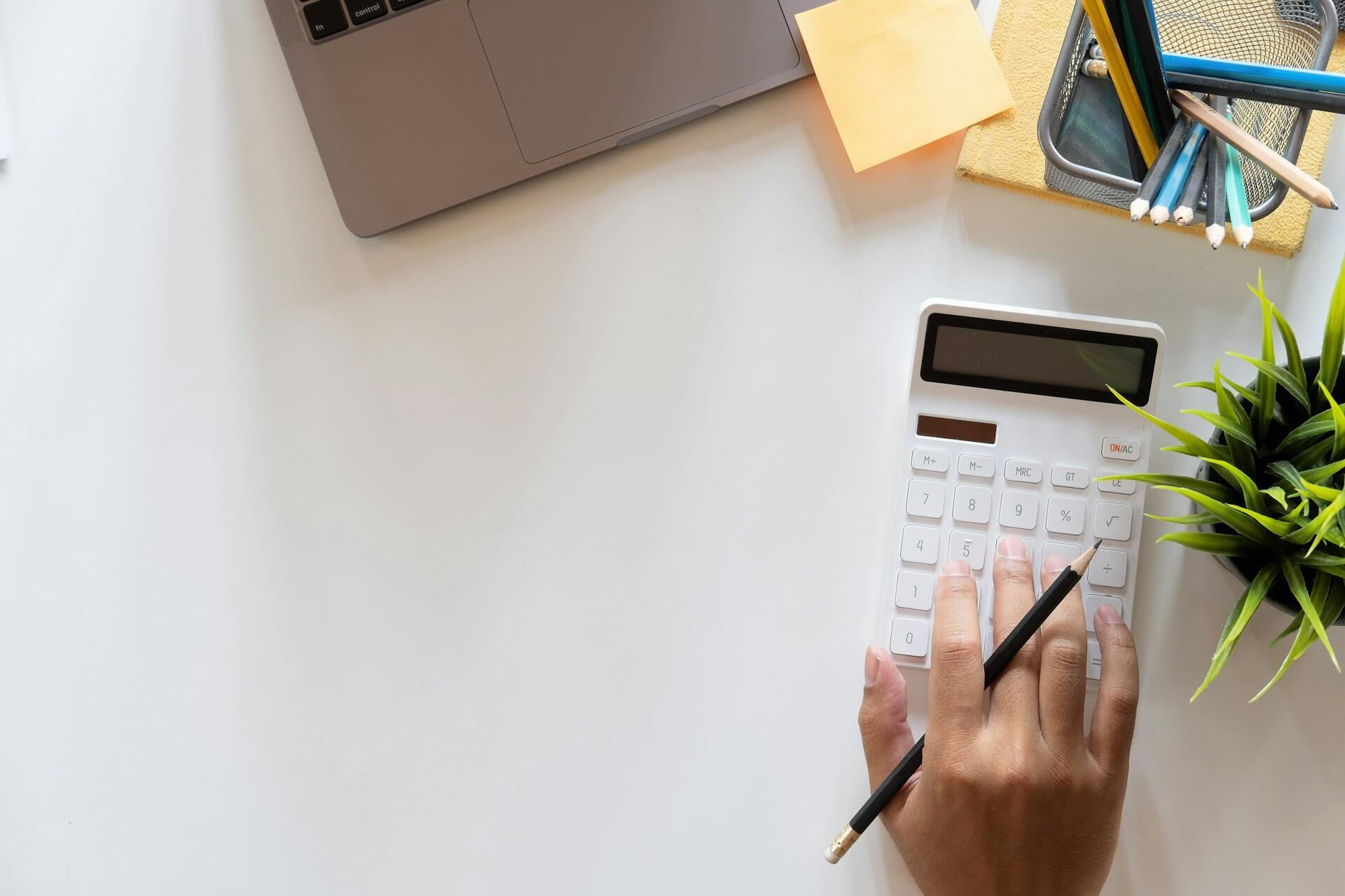 A hand uses a calculator while a laptop, a plant, and stationery are arranged on a clean workspace.