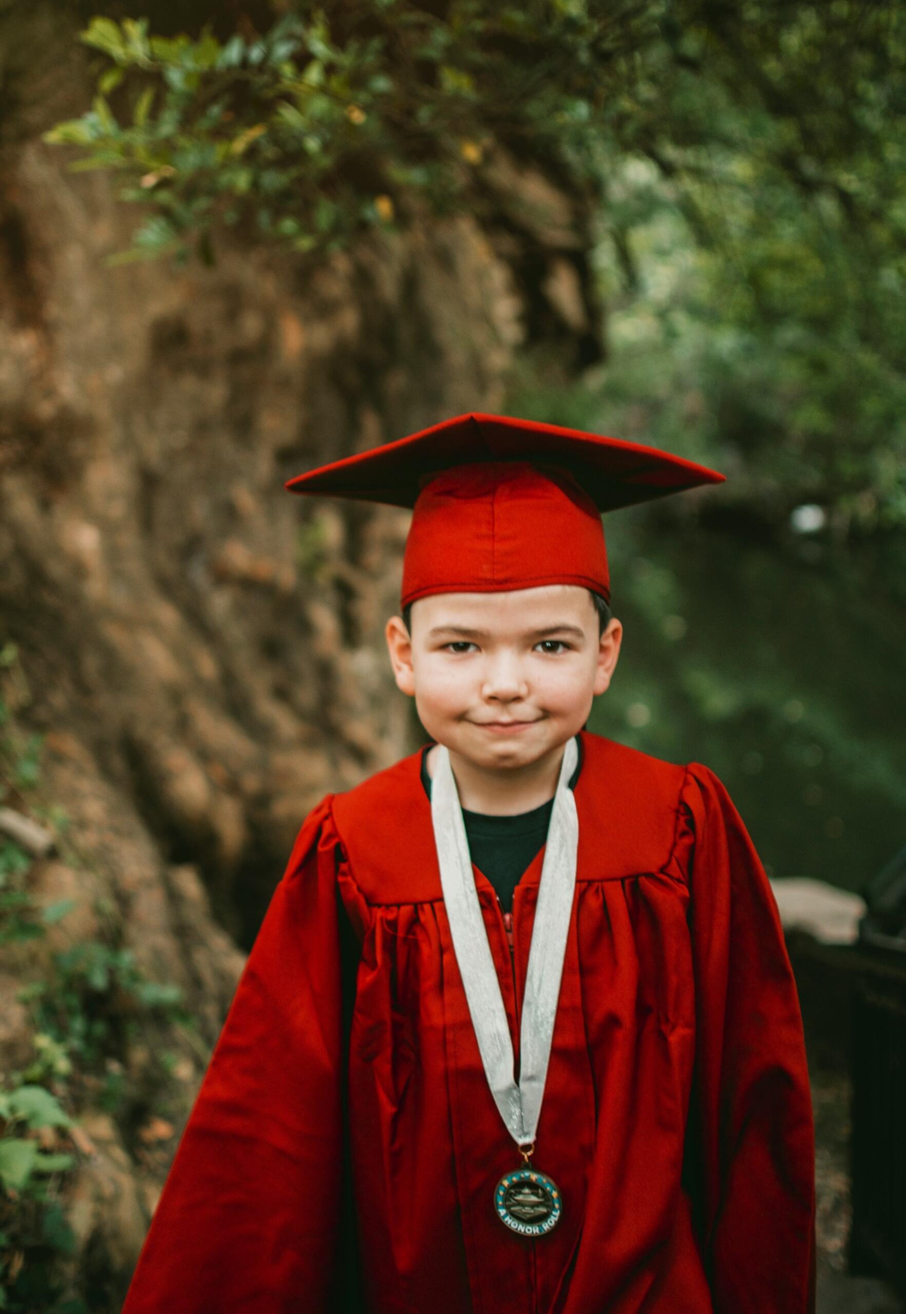 A boy in graduation regalia standing next to a tree. 