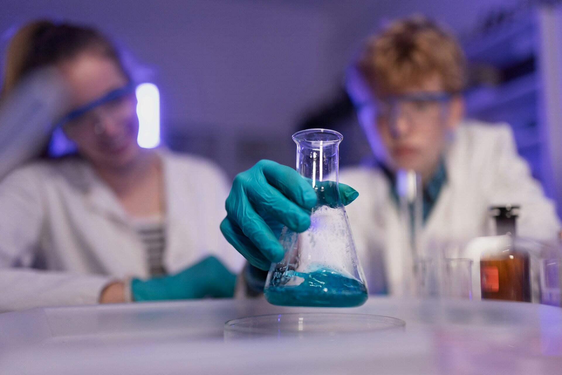A chemist in a lab coat holds a glass flask with bubbling blue liquid.