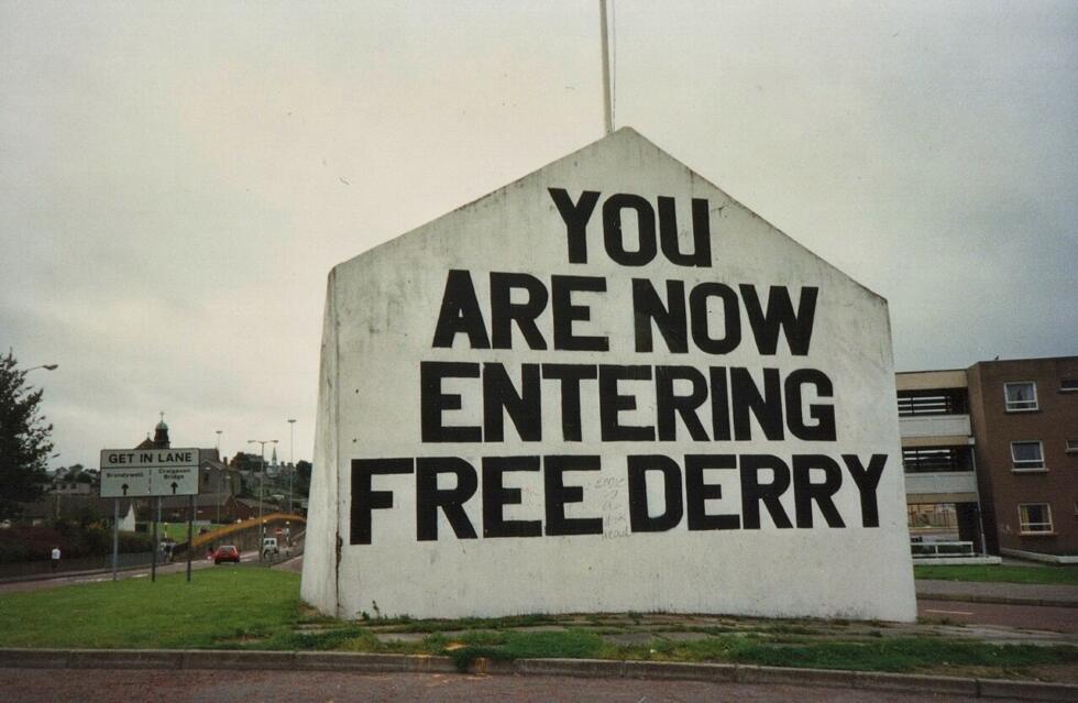 Large white wall with bold black text reading "YOU ARE NOW ENTERING FREE DERRY" beside a road and buildings.