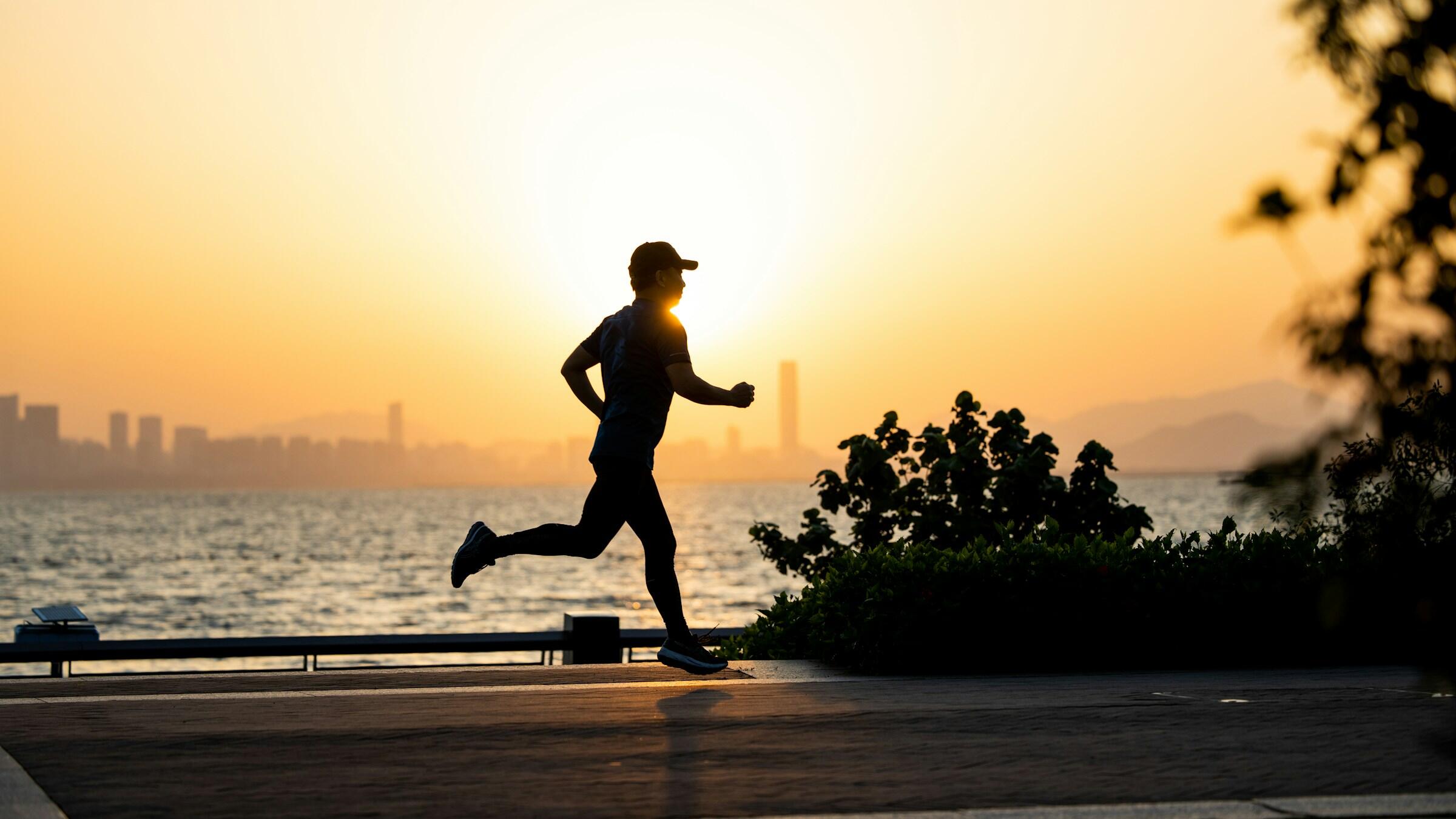 A silhouetted runner jogs along a waterfront promenade at sunset.