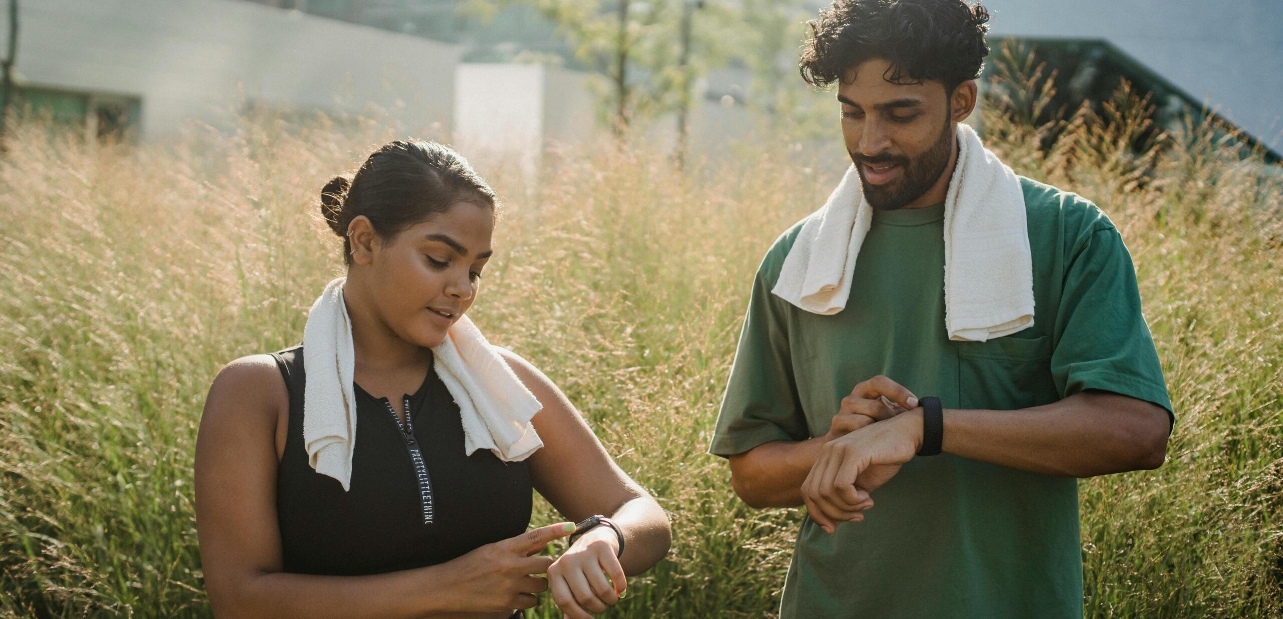 A man and woman outdoors, wearing athletic attire with towels around their necks, checking fitness watches.