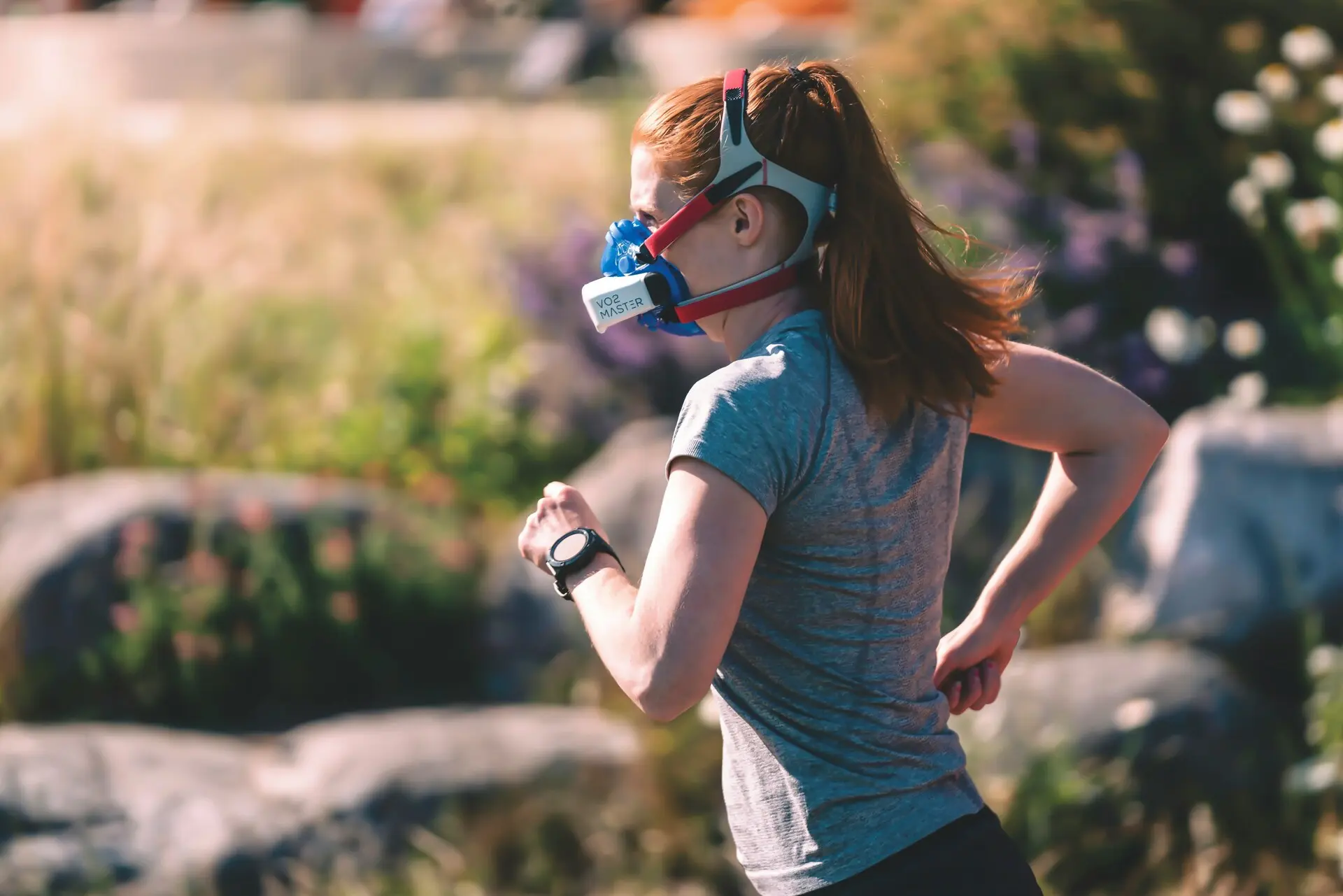 A woman runs outdoors wearing a sports shirt, wristwatch, and a training mask, surrounded by lush greenery and rocks.