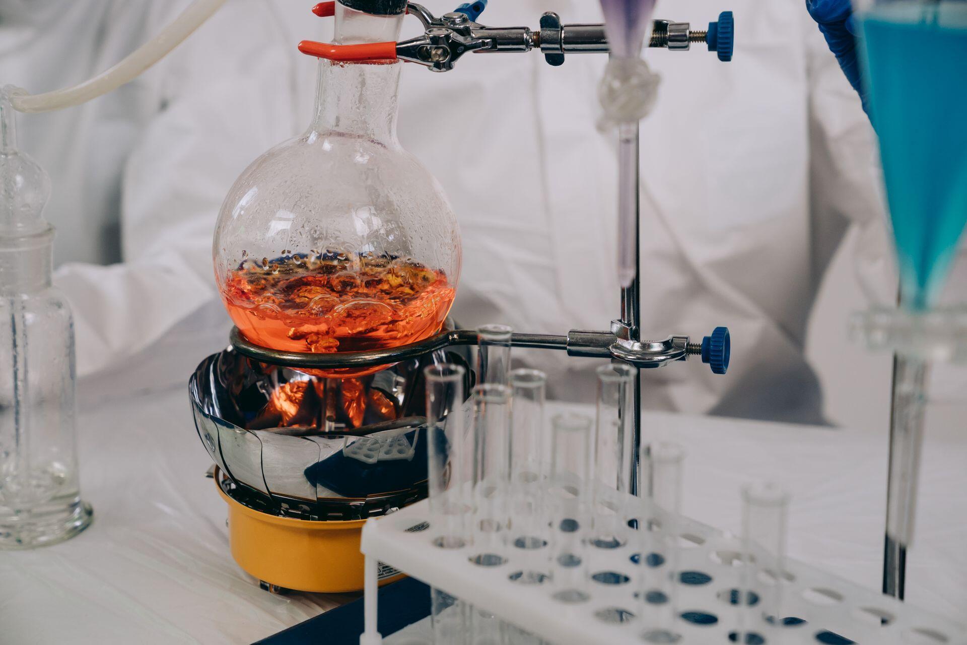 a chemist in a lab coat observing a glass container filled with boiling liquid suspended above a fire in a laboratory
