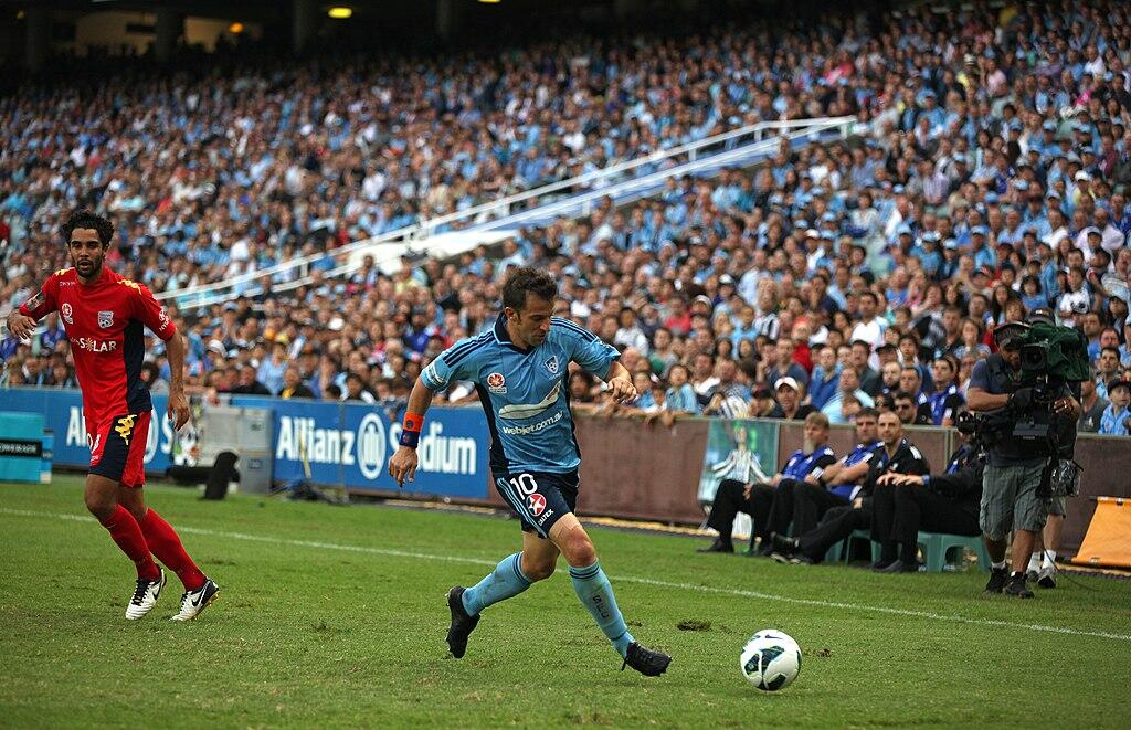Italian soccer player Del Piero in a blue Sydney FC jersey dribbles the ball on the field.