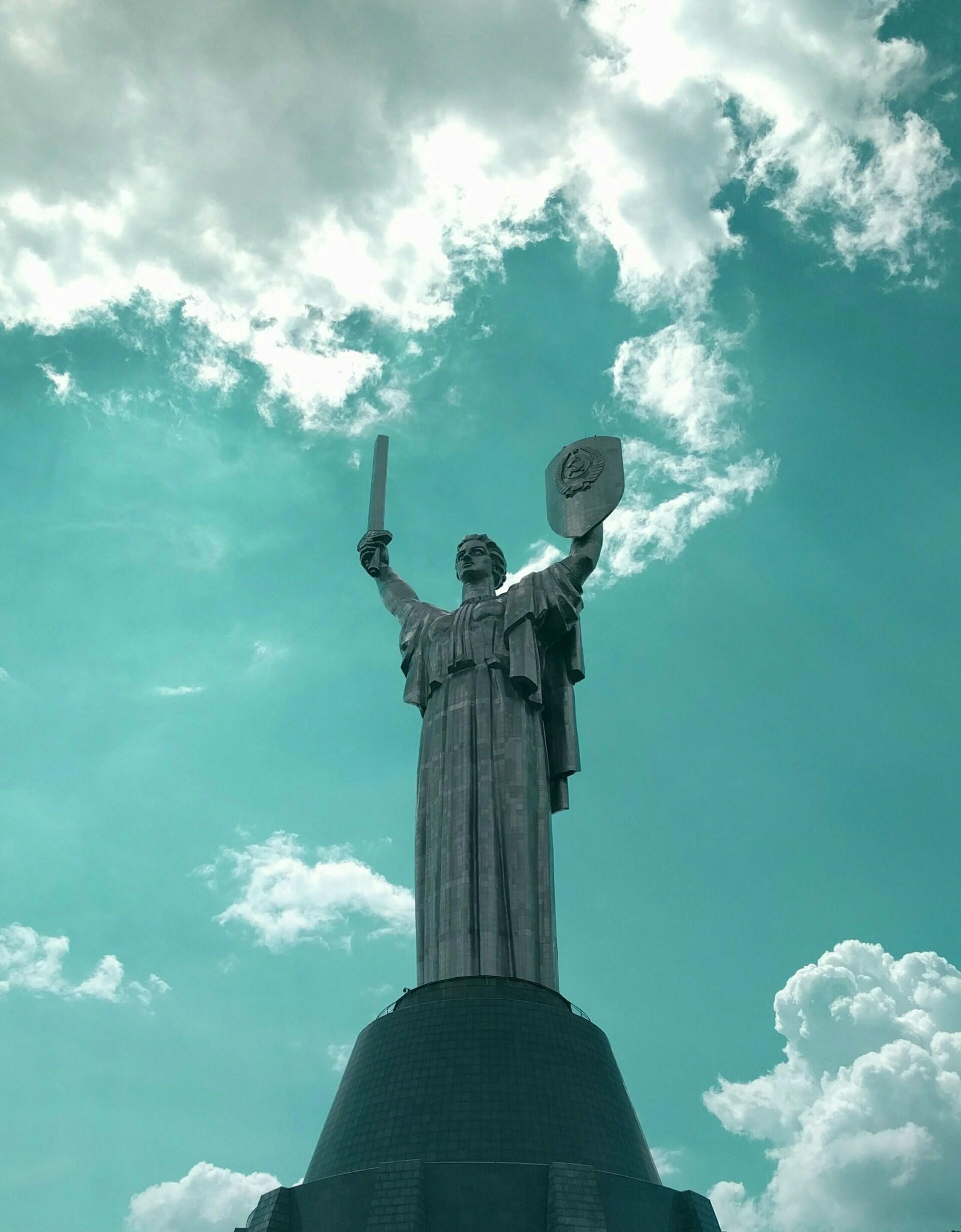 A towering statue of a warrior holding a sword and shield against a turquoise sky with fluffy clouds.