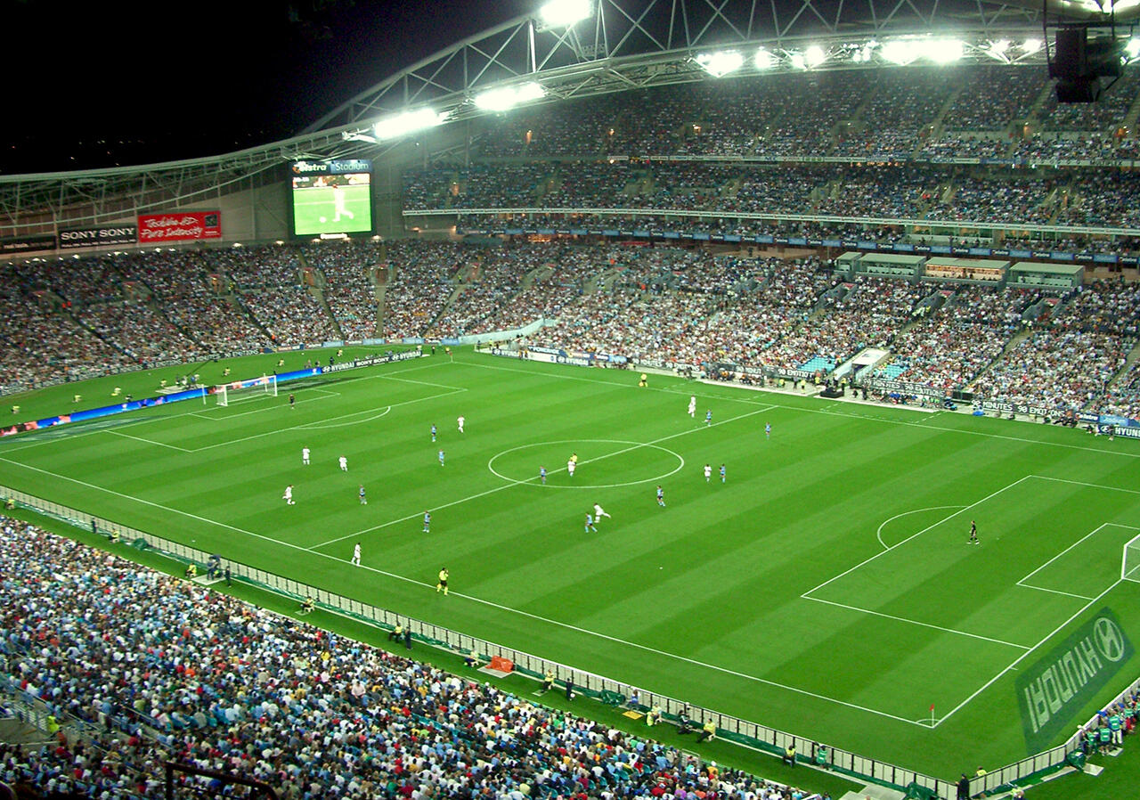 A packed Stadium Australia under bright lights during a soccer match between Sydney FC and LA Galaxy.