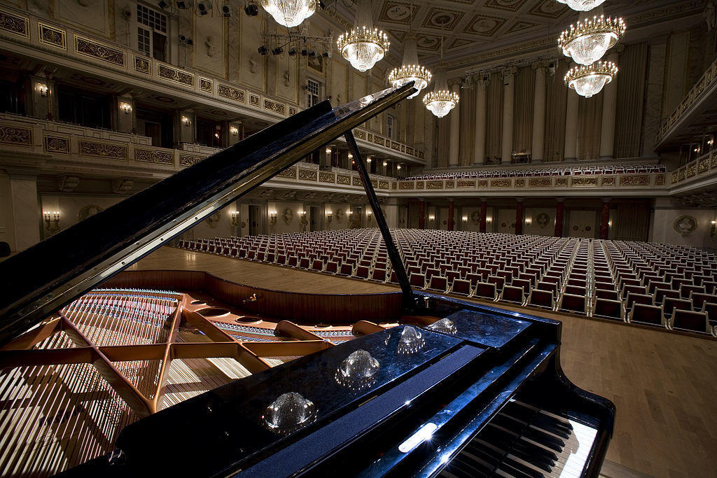 A grand piano with open lid in the foreground, overlooking an elegant concert hall filled with empty red chairs and chandeliers.