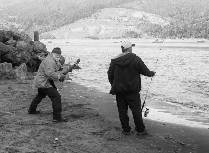 A bearded man plays guitar on the bank of a river - next to a fisherman.
