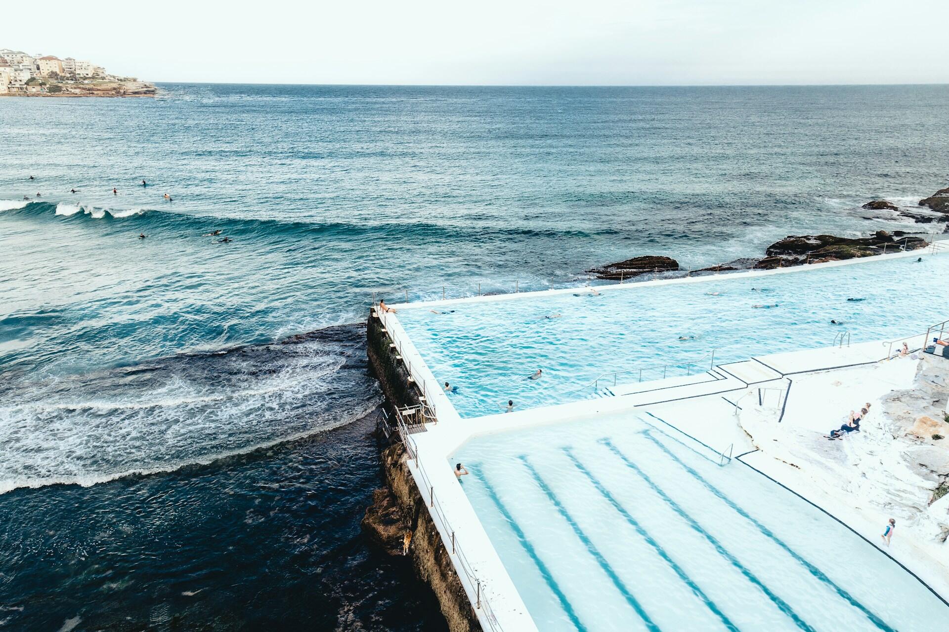 Aerial view of a rectangular ocean pool by a rocky shore, with swimmers in the water and surfers riding waves in the background.