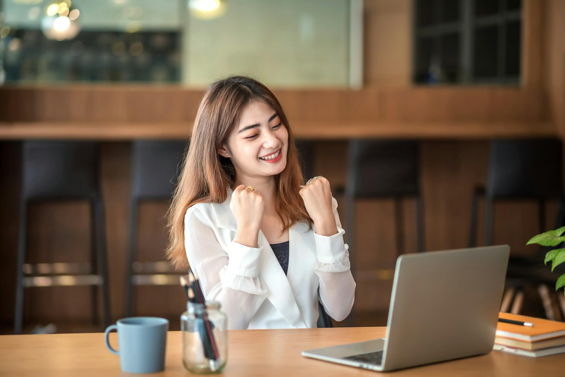 A teacher in a white shirt raises their fists in at a desk with a laptop;