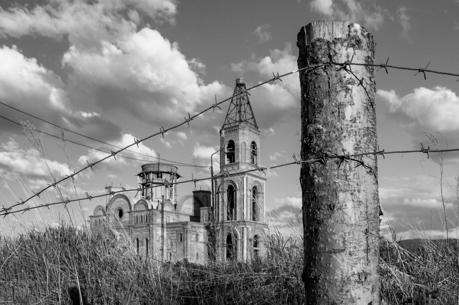 A black and white image of a dilapidated church surrounded by tall grass, with barbed wire fencing.