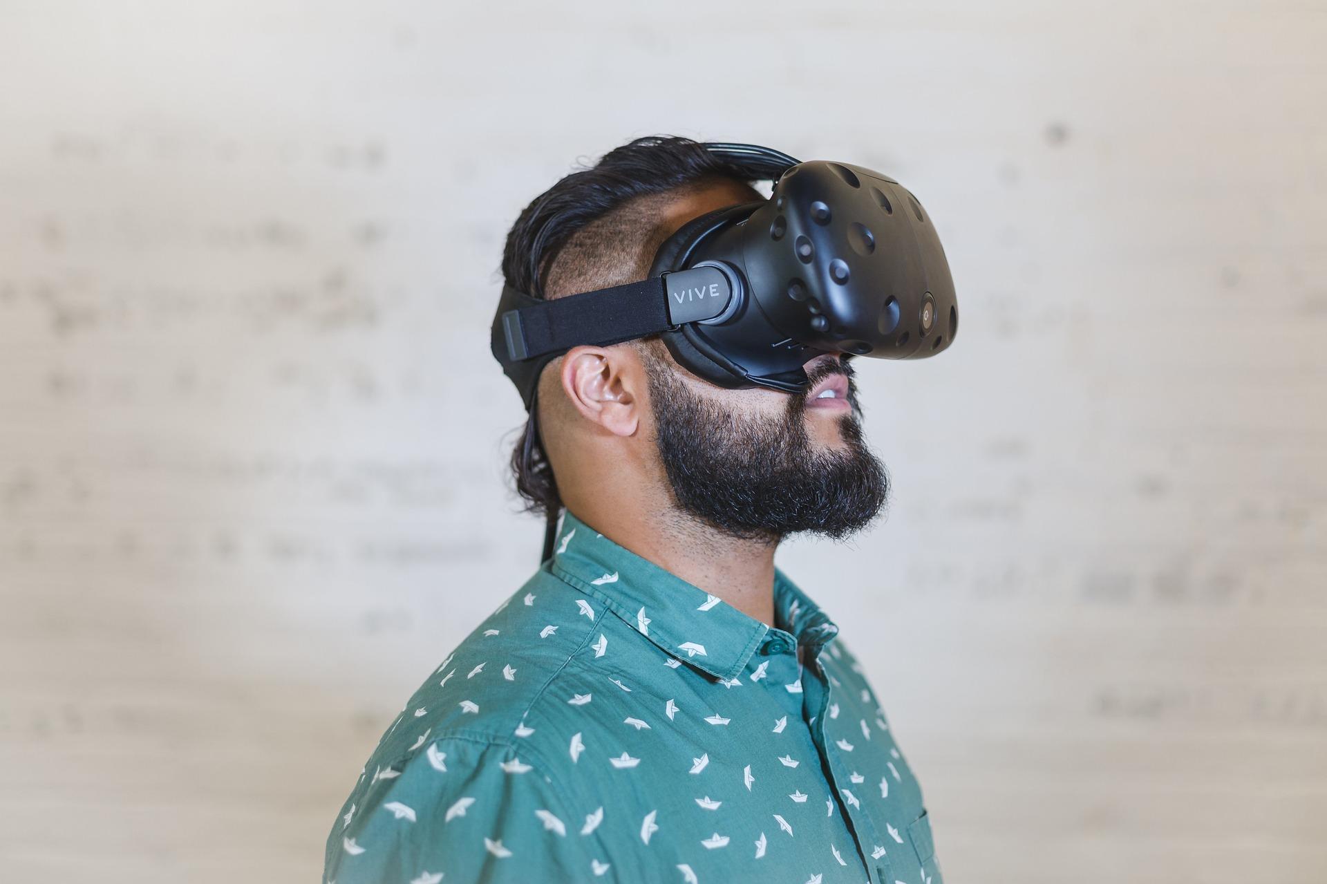 A bearded man in a print green and white shirt wears a virtual reality headset, his head tilted slightly upwards.