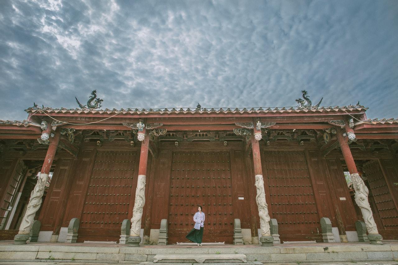 A person in traditional attire stands before a grand wooden building with intricate carvings and a dramatic cloudy sky above.