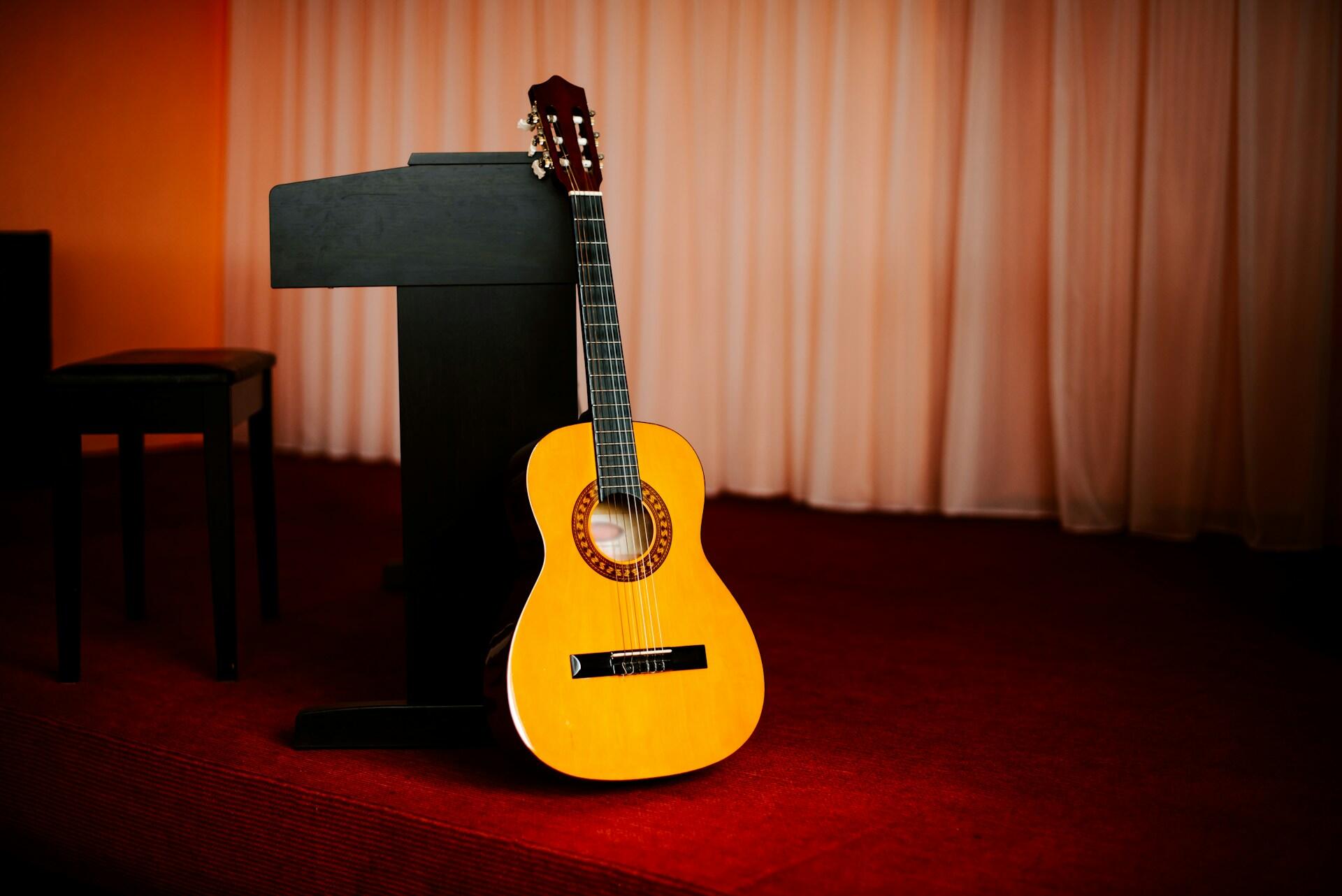 An acoustic guitar leans against a black music stand on a red carpeted stage.