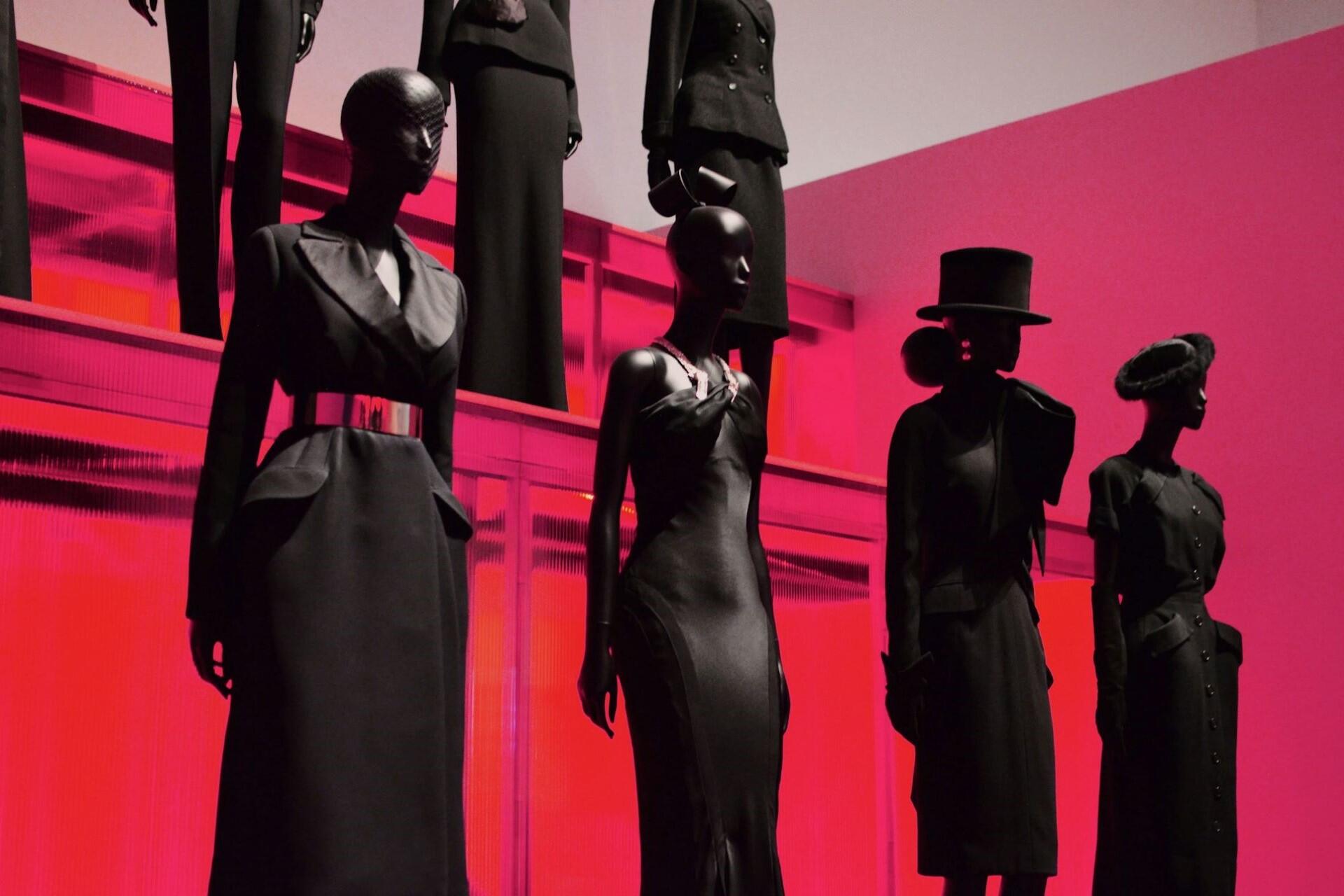 Mannequins wearing black garments displayed in a fashion exhibition with red background lighting