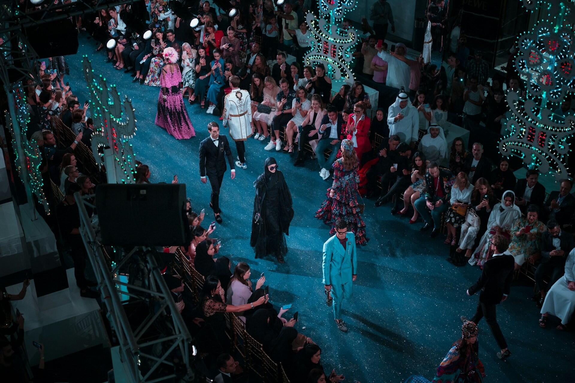 Models walking on a fashion runway surrounded by seated audience members and decorative lighting