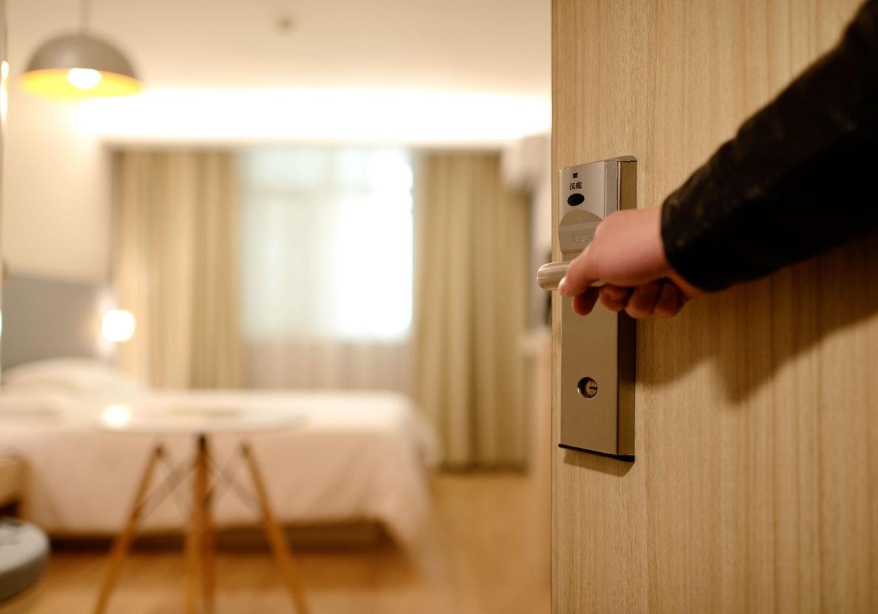 A hand reaches for a door handle, opening into a softly lit hotel room with a bed and curtains in the background.