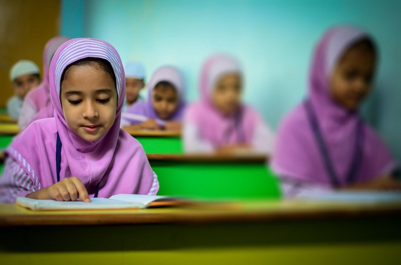 A classroom scene featuring students in pink hijabs, attentively studying with books on their desks. Brightly painted green accents visible.