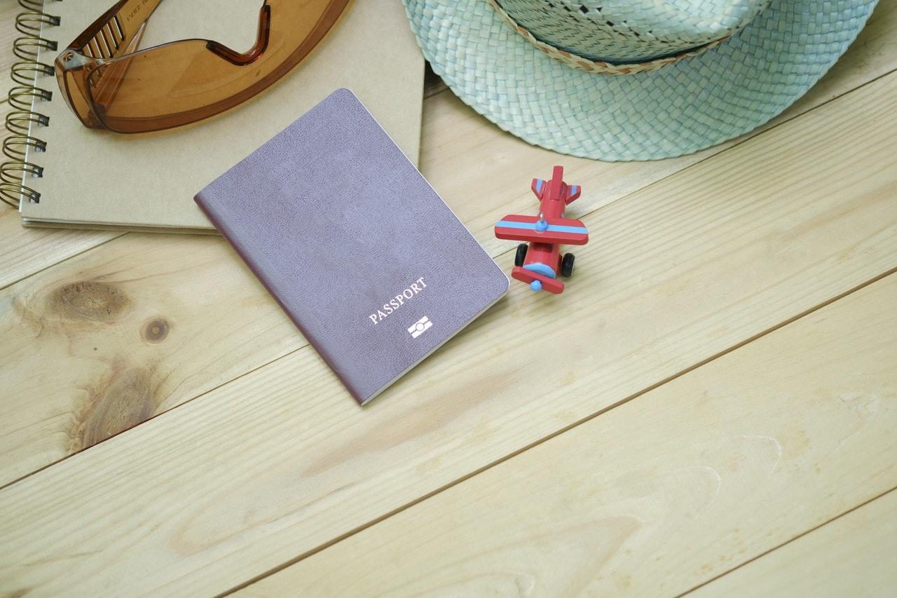 A brown passport, toy airplane, sketchbook, and sunglasses are arranged on a wooden surface, accompanied by a straw hat.