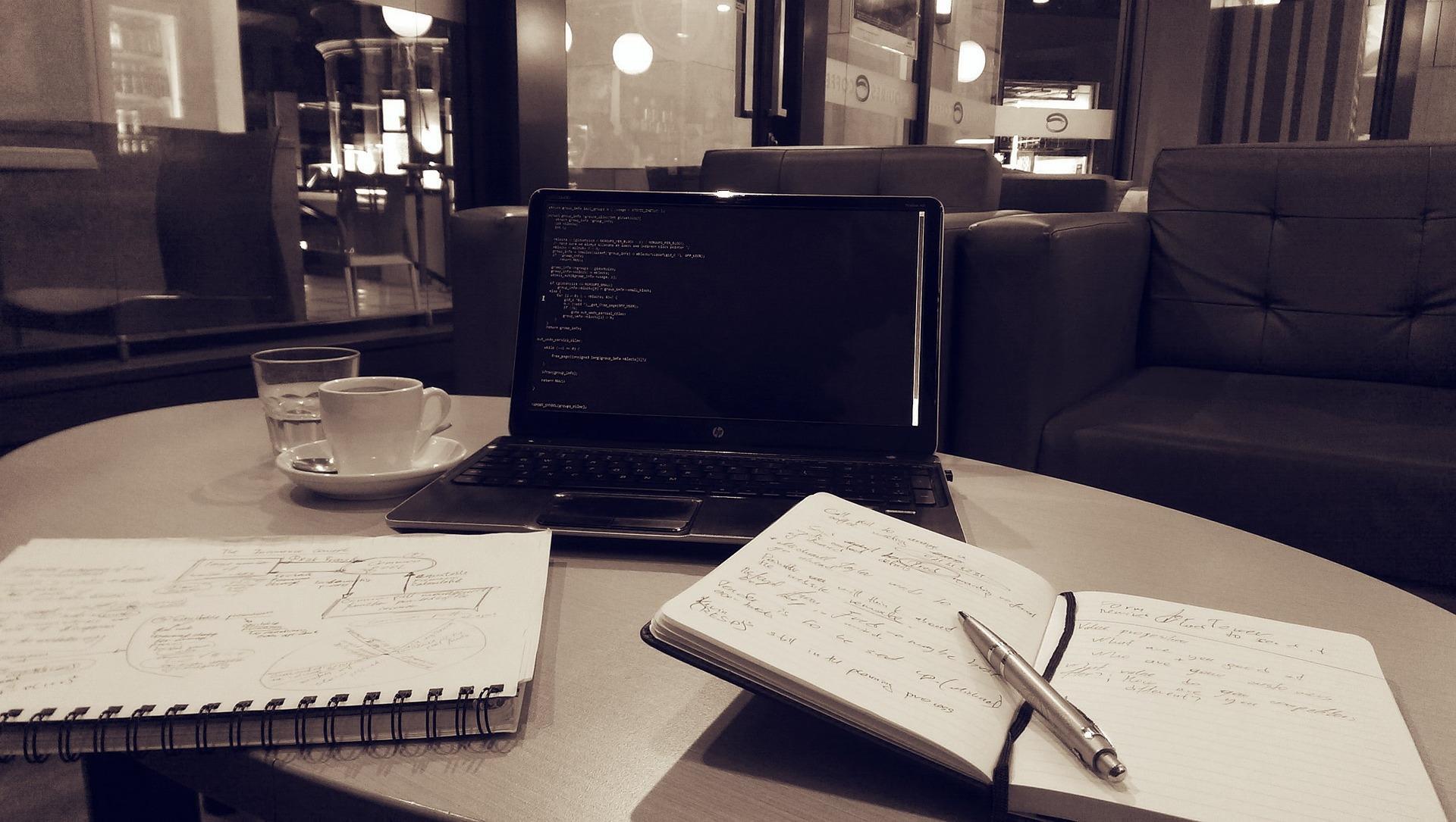 An open laptop computer whose dark-mode screen displays colourful lines of code rests on a round, white-topped table, behind an open notebook with a pen laying across its spine and a spiral notebook lying lengthwise. Next to the computer is a white up and saucer and, behind it, a glass of water.