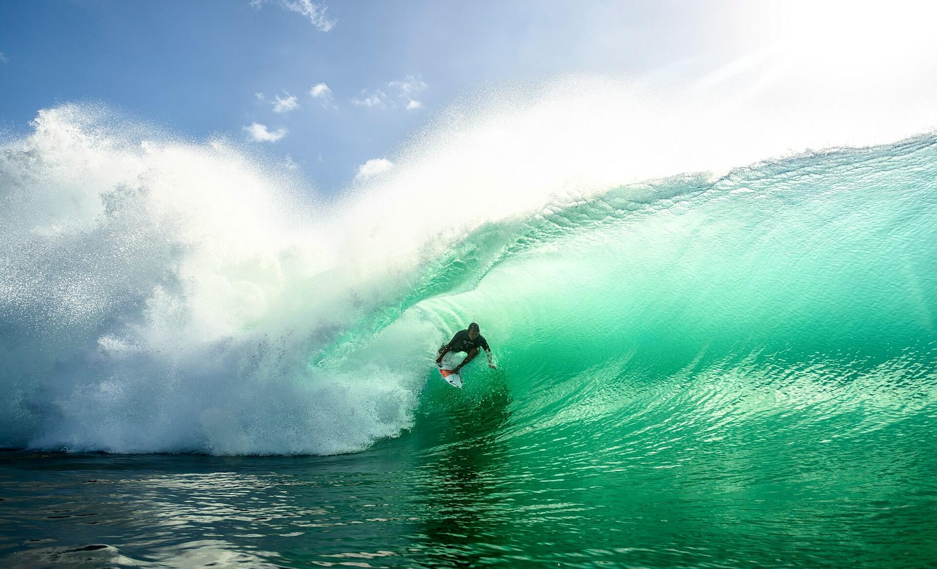 A surfer navigates a large, turquoise wave under a bright blue sky, creating a dynamic scene of surfing adventure.