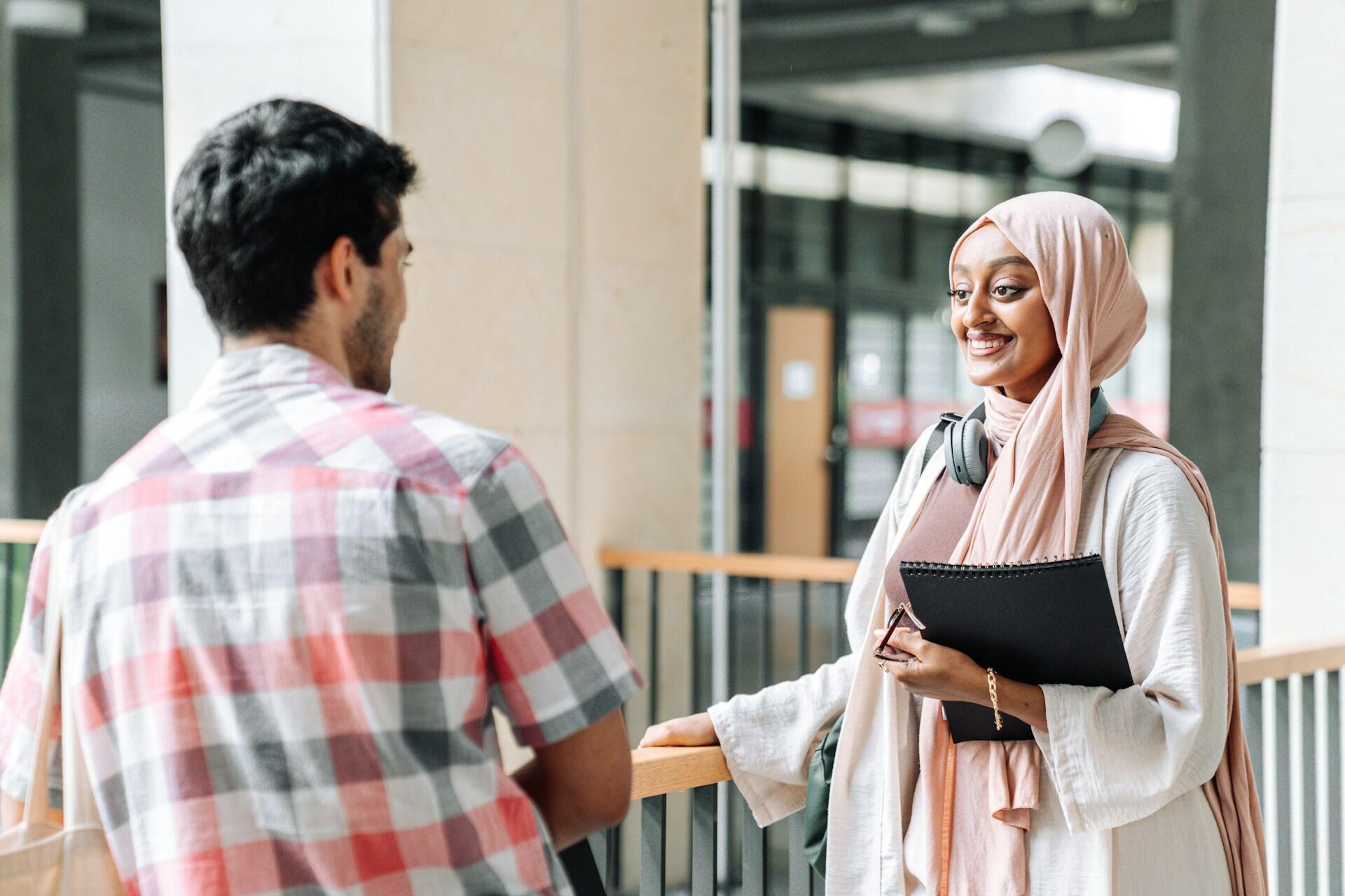 A woman in a light hijab holds a notebook, engaging in conversation with a man.