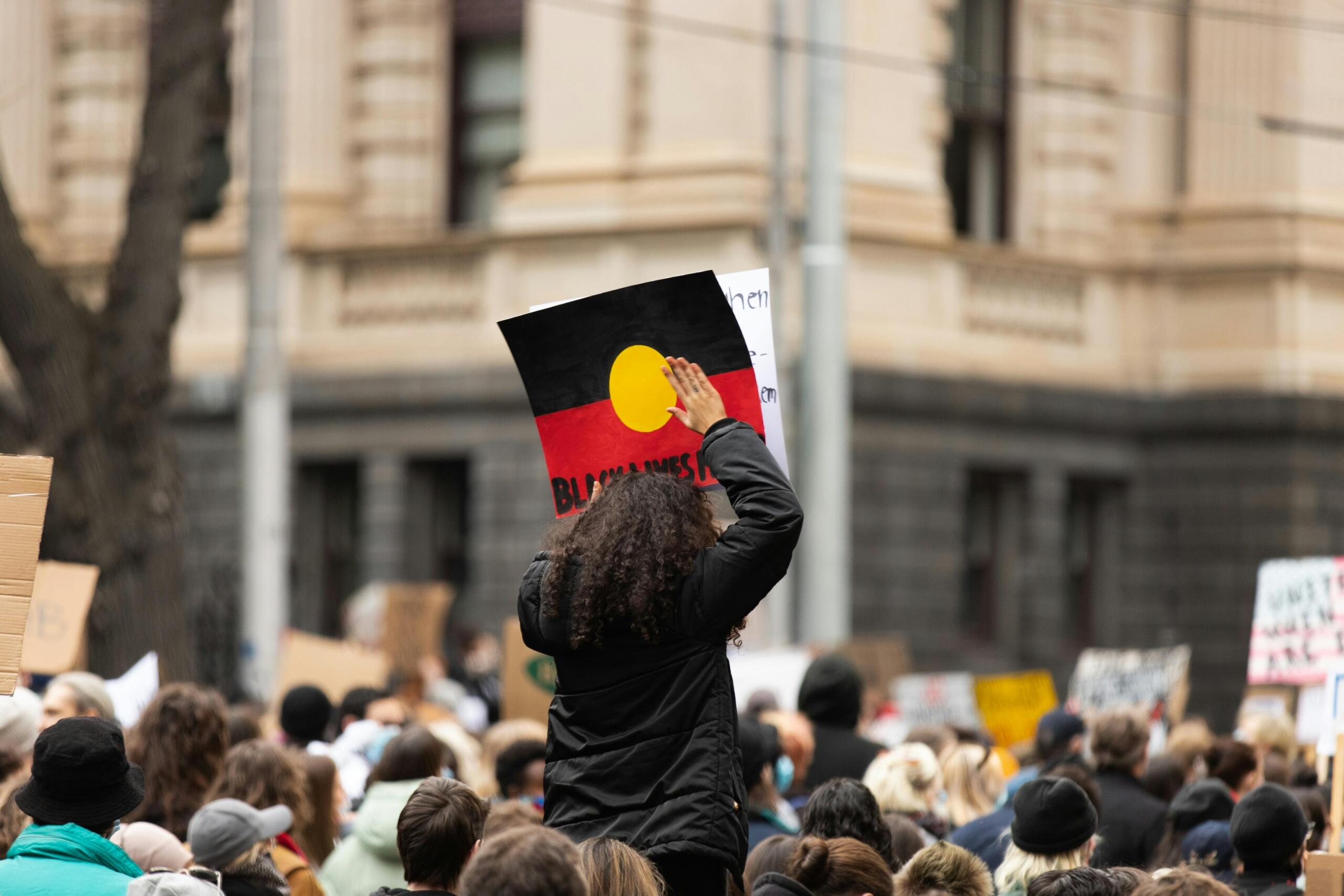 A rally scene with a crowd holding signs; one person raises an Australian Aboriginal flag.