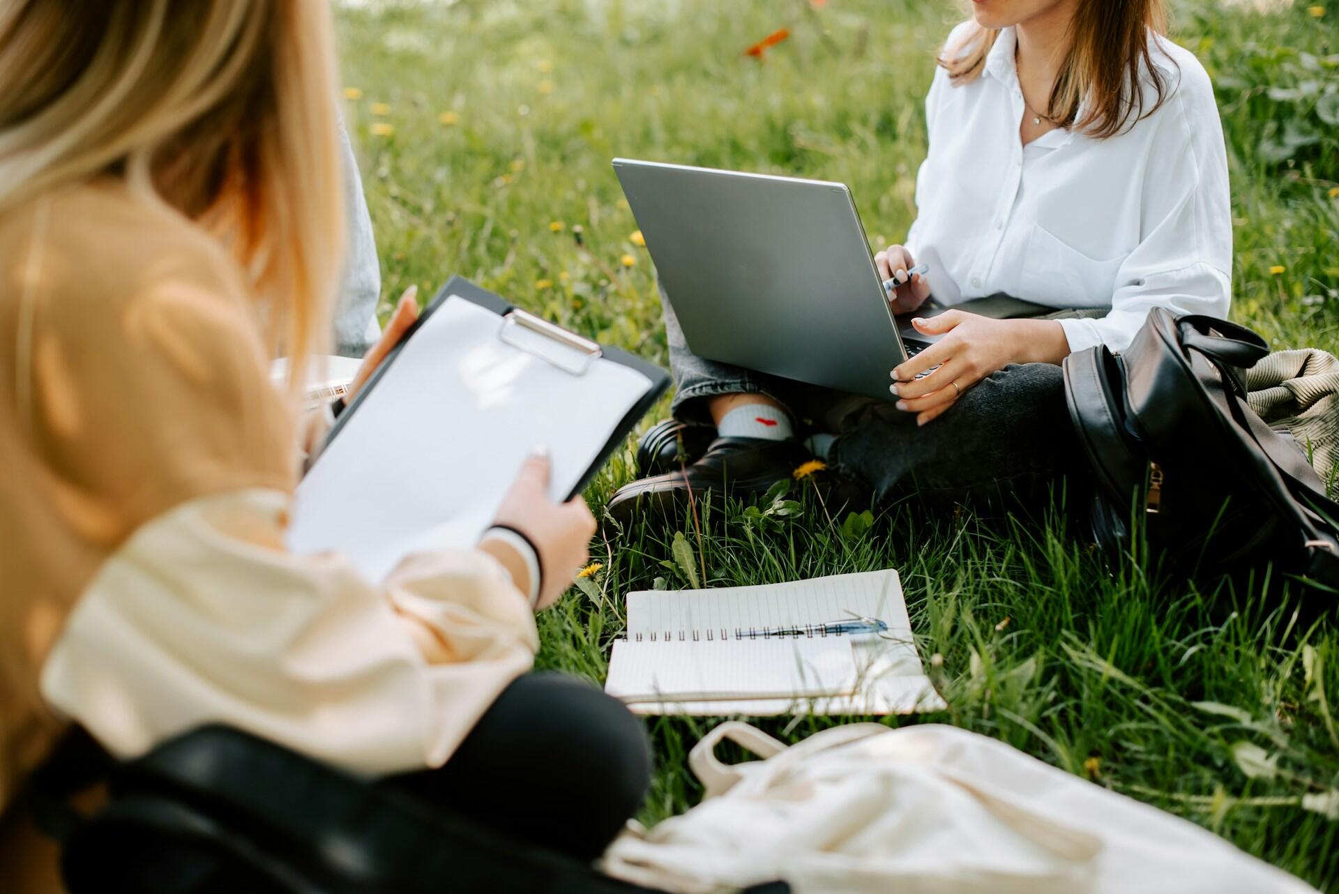 Two people sit on the grass, one with an open computer and the other with a clipboard.