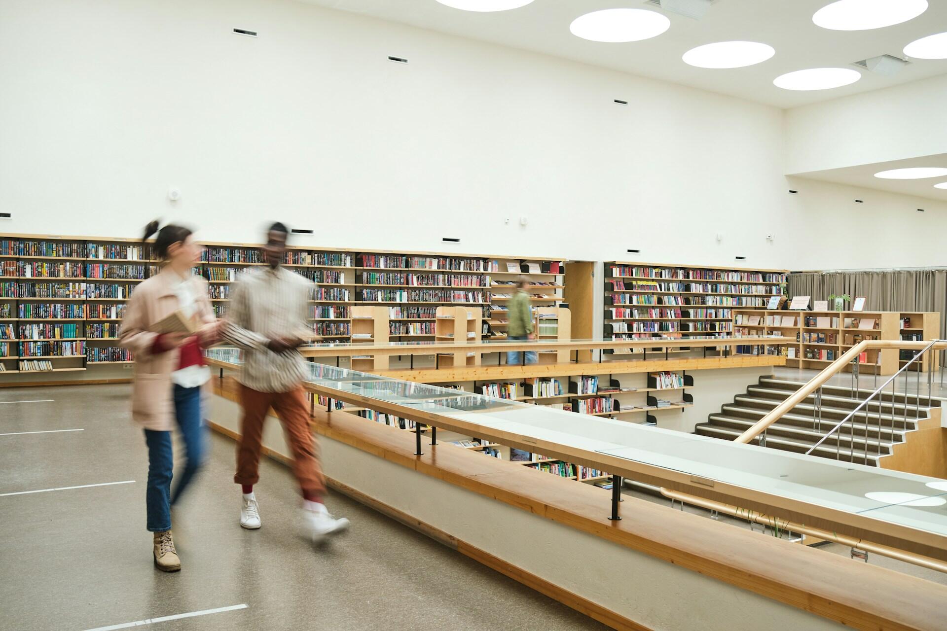 Two people walk through a modern library, surrounded by bookshelves.
