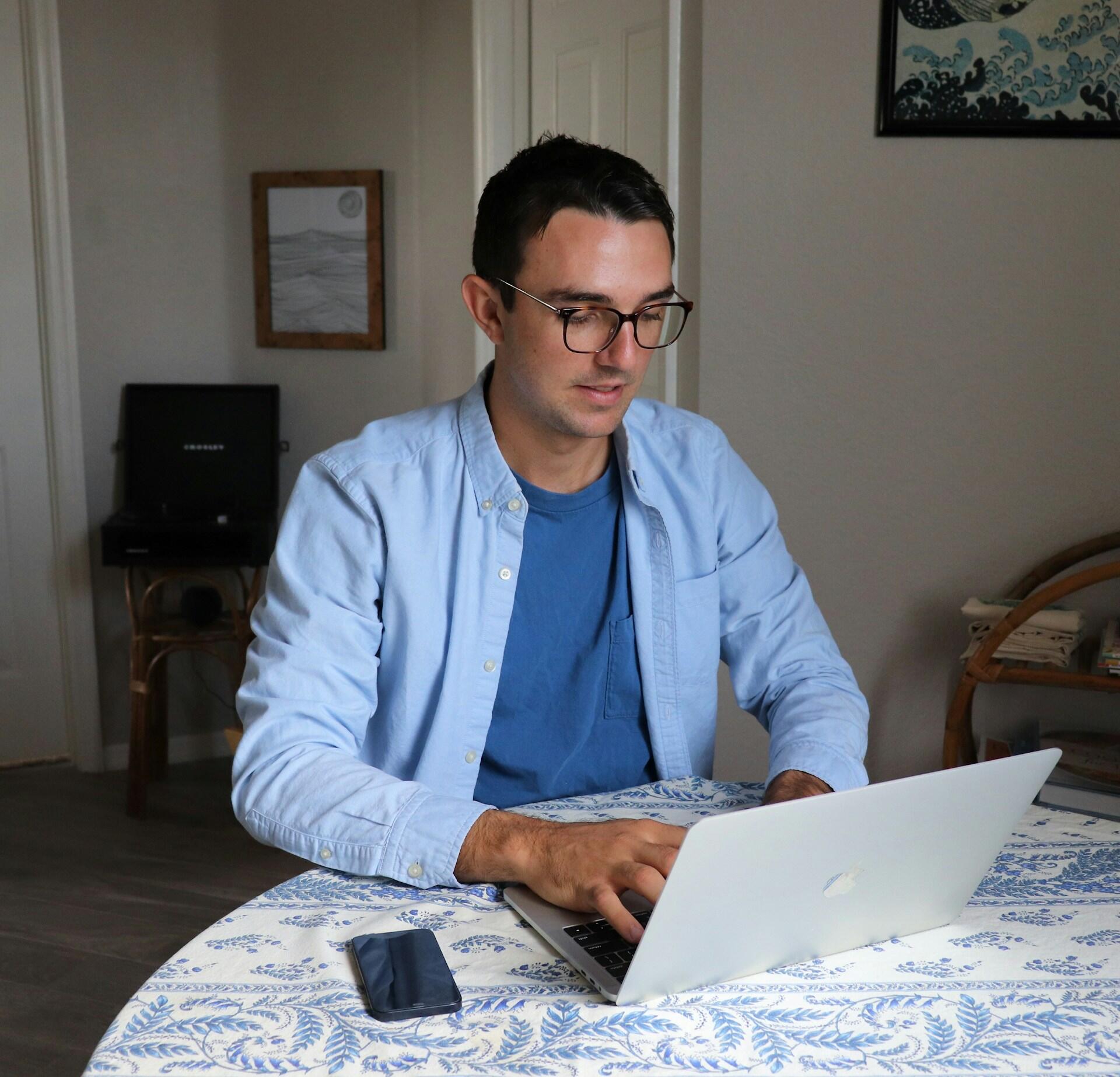A man wearing blue sits at a table using a computer.