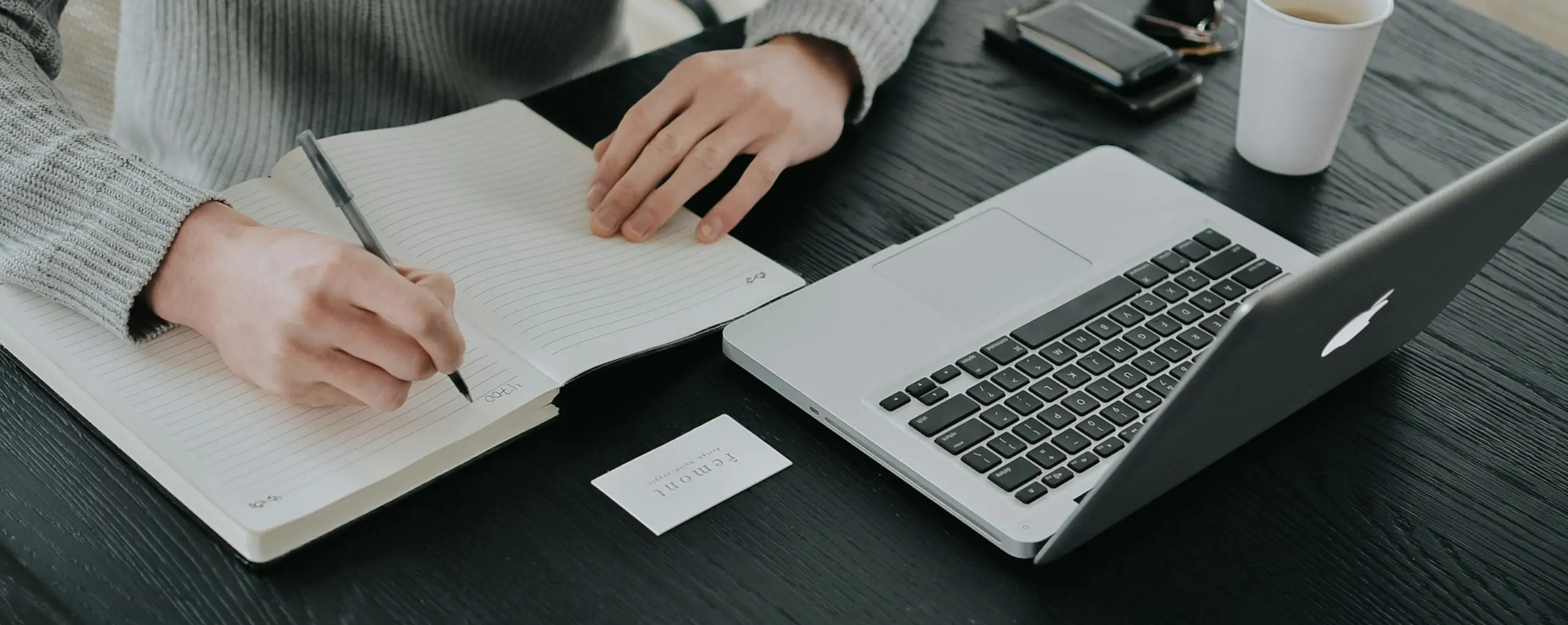 A person writes in a notebook while seated at a desk with a laptop.