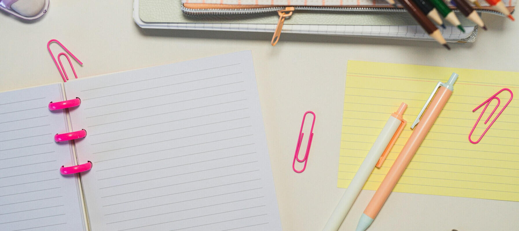 Flat lay of white lined notebook, yellow index card, two retractable pens, pink paperclips, colored pencils, and a pencil case on a light desk.