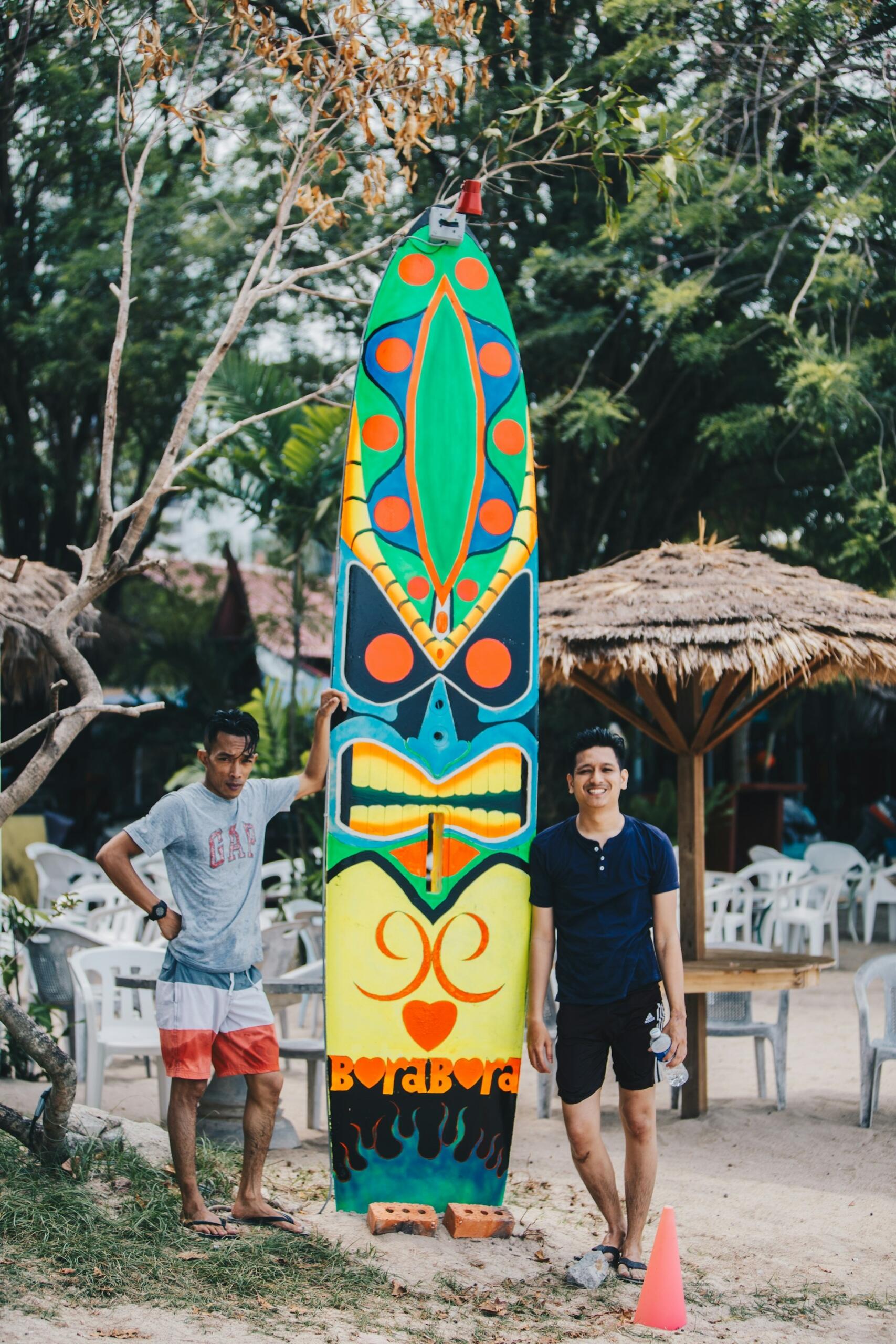 Two people standing next to a tall painted surfboard on a beach.