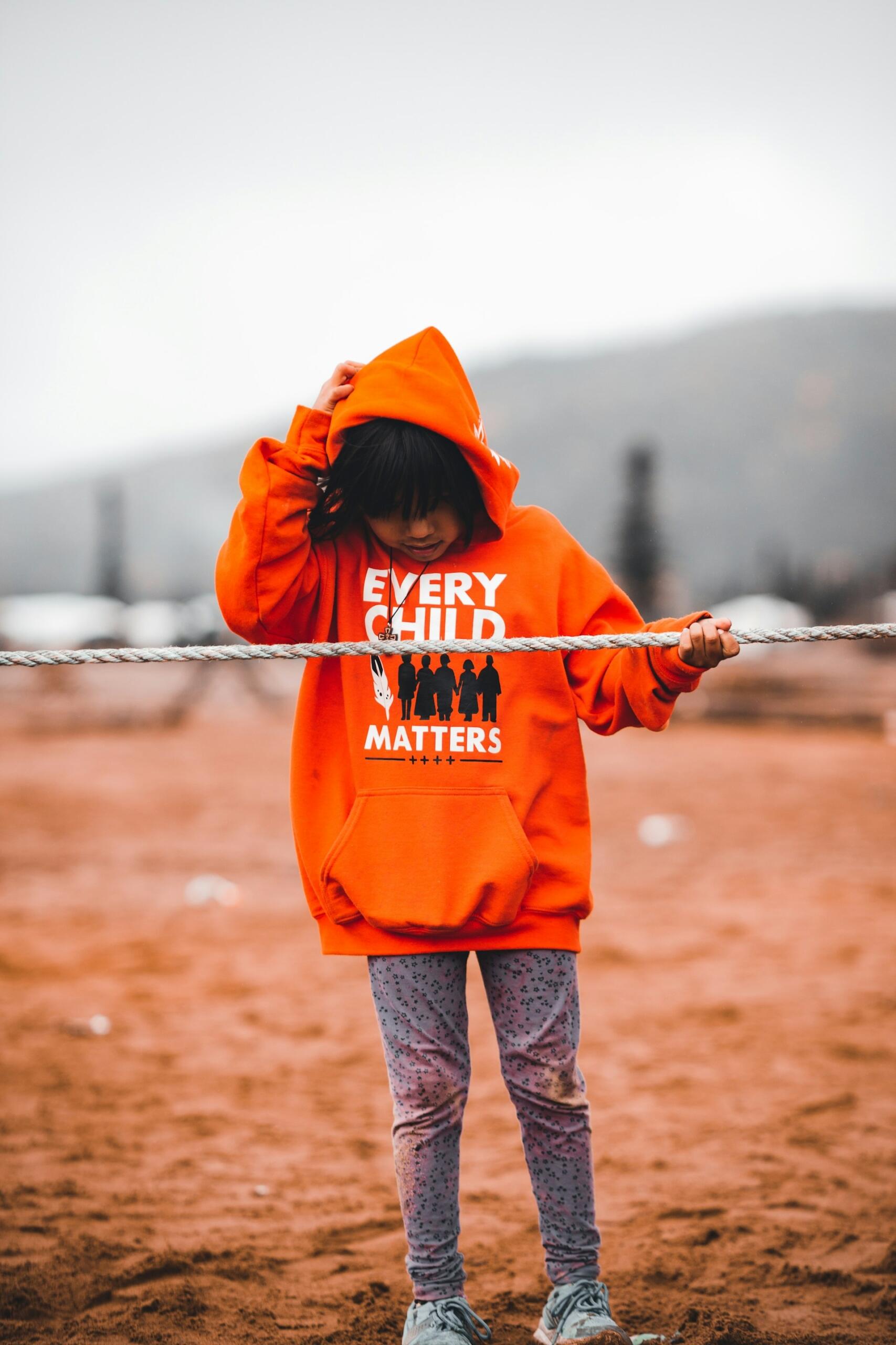 A child wearing an orange hoodie standing next to a fence.
