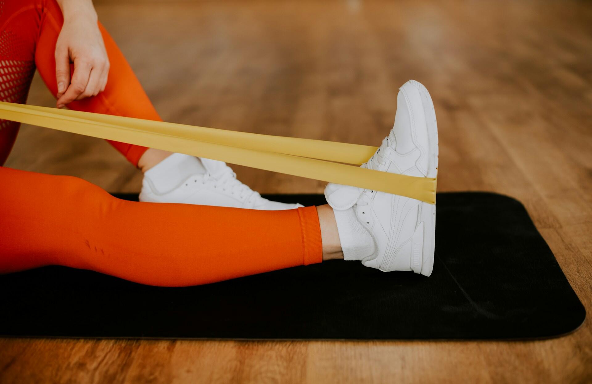 A person in bright orange leggings uses a yellow resistance band while seated on a black mat.
