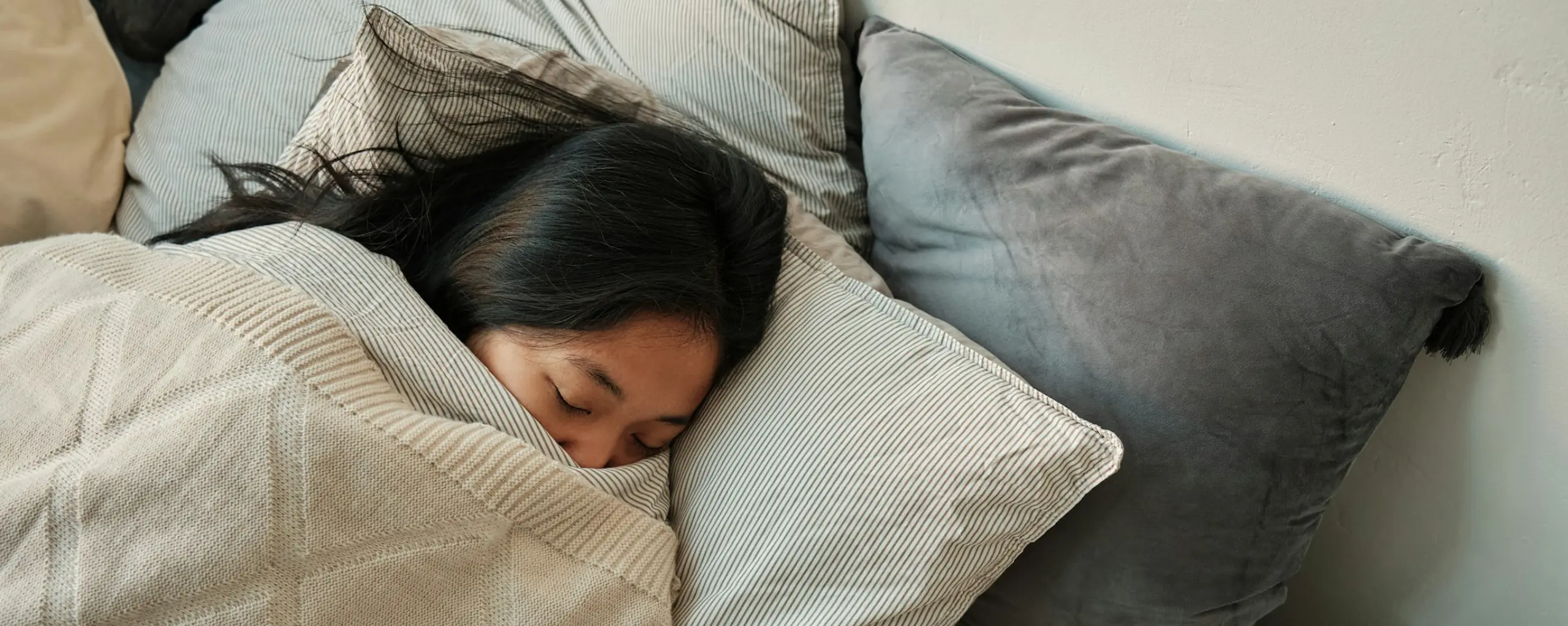 A person sleeping under a knitted blanket amid soft pillows.