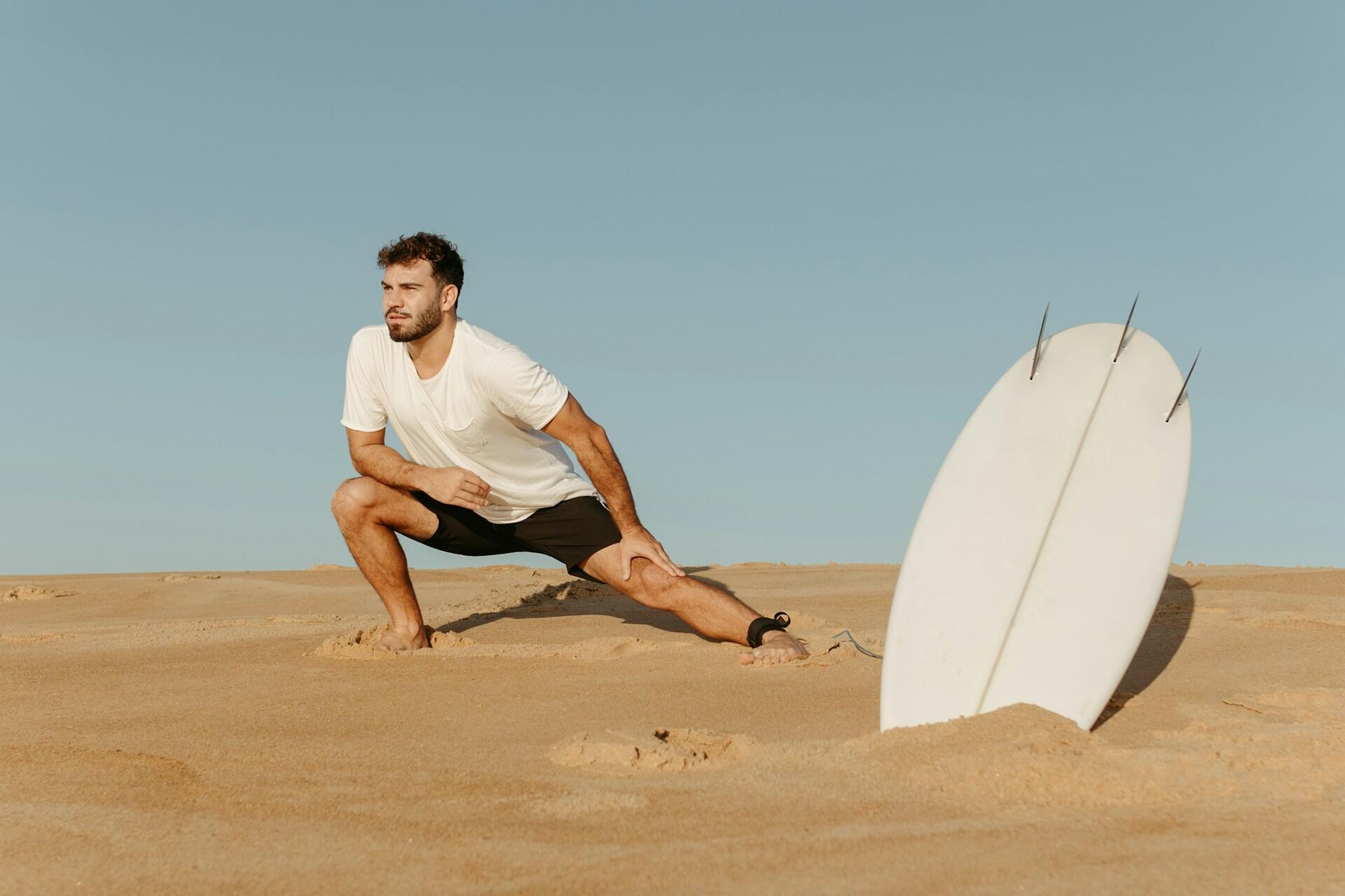 A man in a white shirt and black shorts stretches on a sandy beach next to a surfboard.