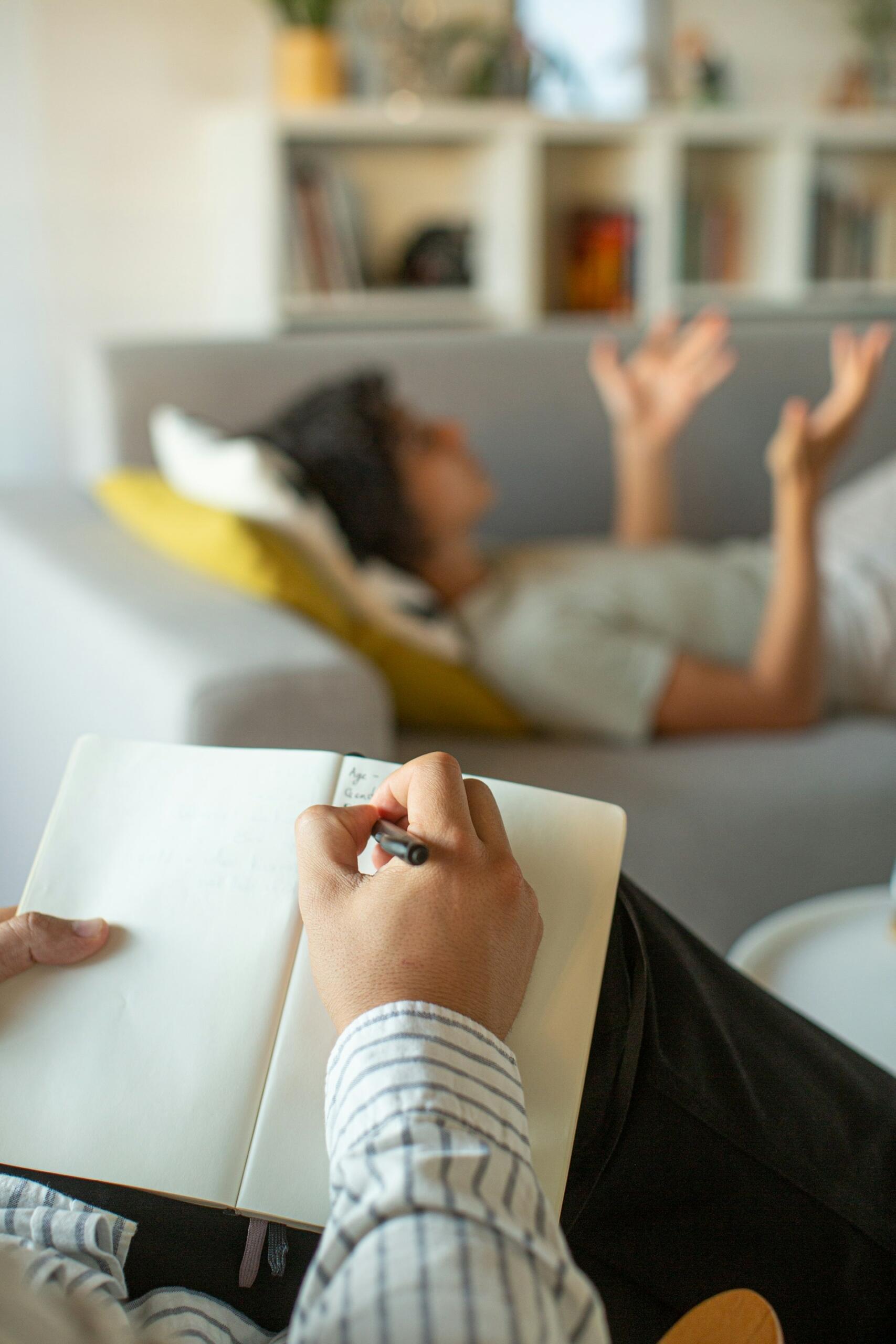 A person sits with an open notebook an pen in hand while another lies on a nearby couch.