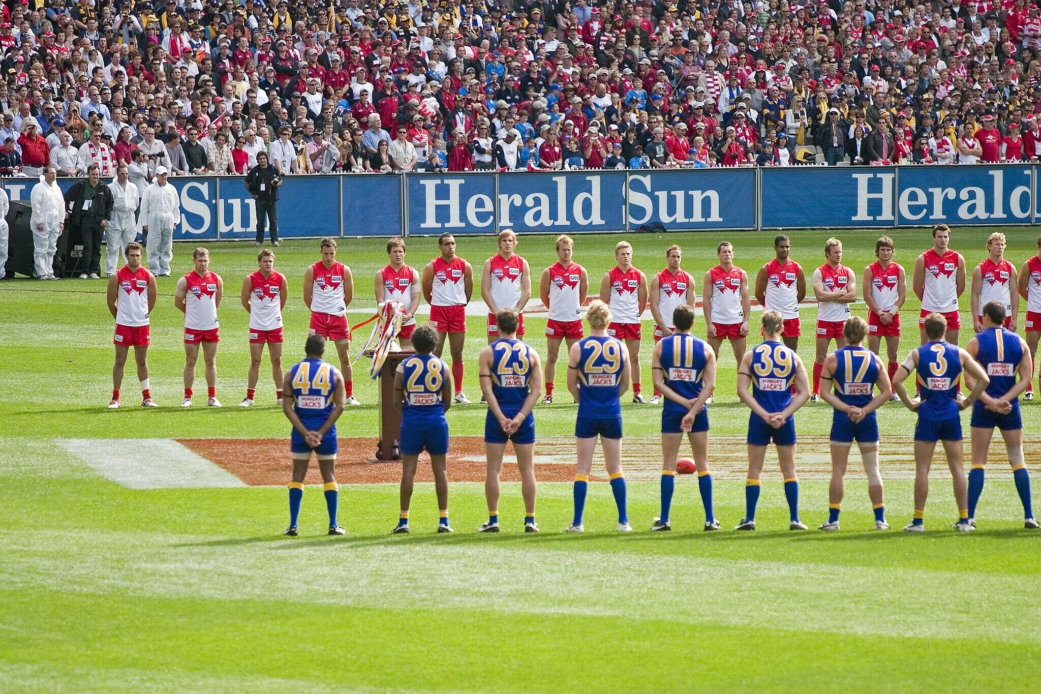 Aussie rules football teams lined up on the field in front of a large crowd.